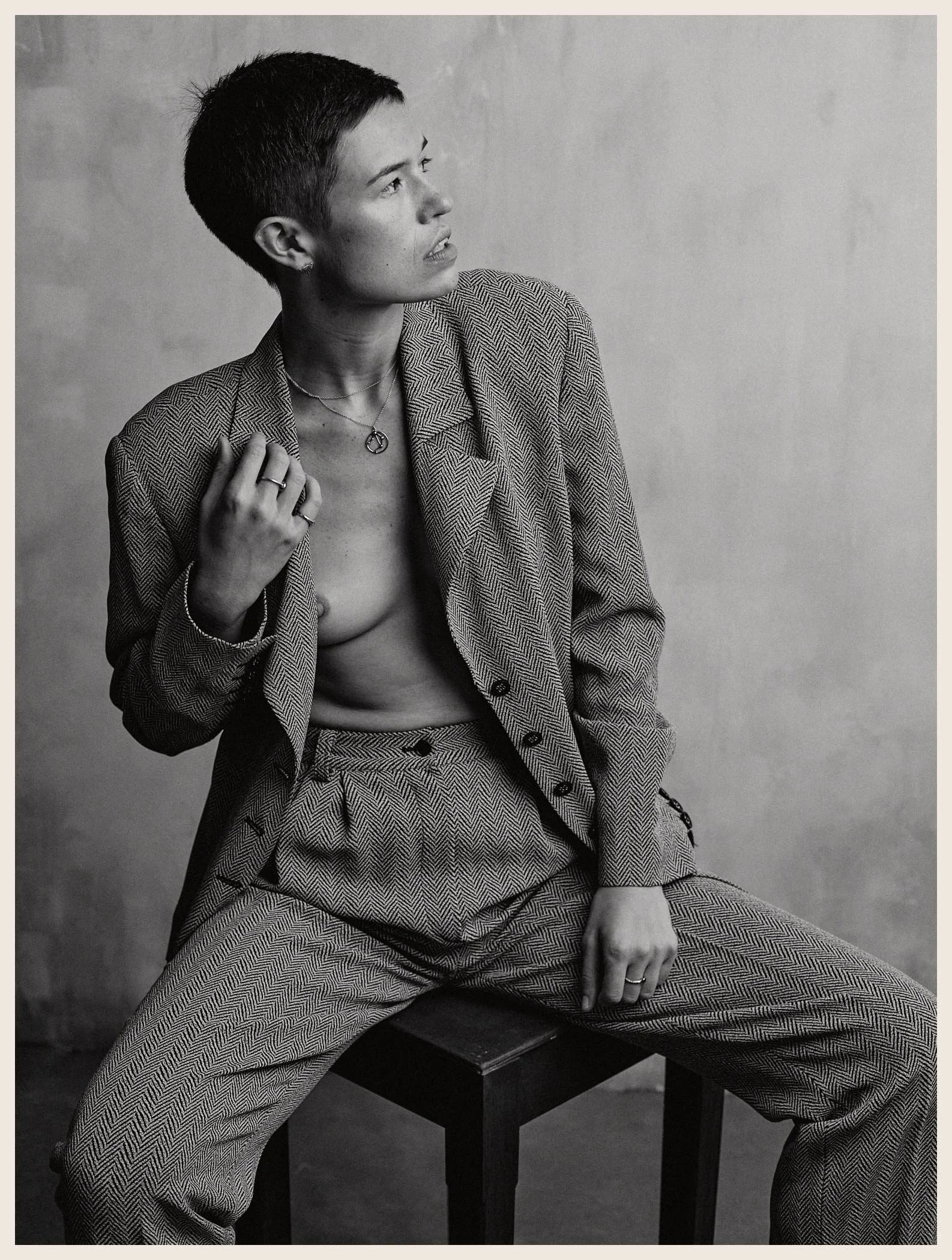 Black and white portrait of a woman with short hair, wearing a herringbone suit jacket and pants, seated on a stool, with one hand on her chest and the other on her knee, looking sideways.