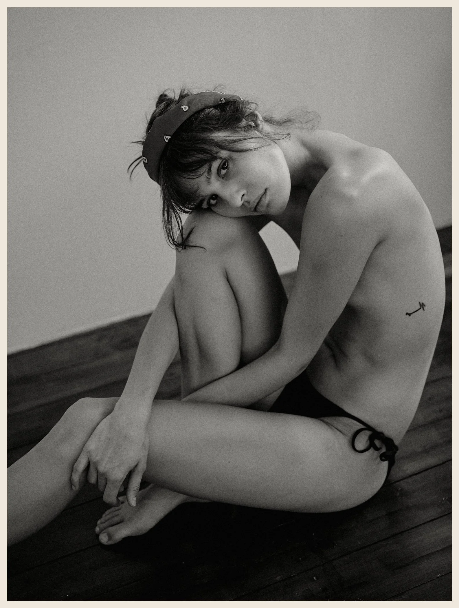 A young woman with a headband, sitting on a wooden floor, hugging her knees, looking at the camera, with a small tattoo on her side, in black and white.