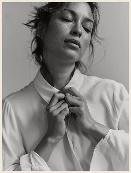 A black and white photo of a woman with her eyes closed, holding her collar with both hands, wearing a loose white shirt.