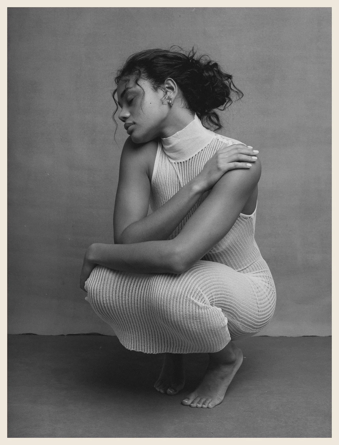 Black and white photo of a woman crouching with her eyes closed, right hand on her left shoulder, wearing a sleeveless, high-neck, striped dress, barefoot, against a plain backdrop.
