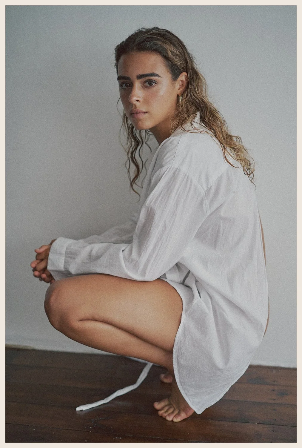 A young woman with curly hair, wearing a white long-sleeved shirt, squatting on a wooden floor against a plain background.