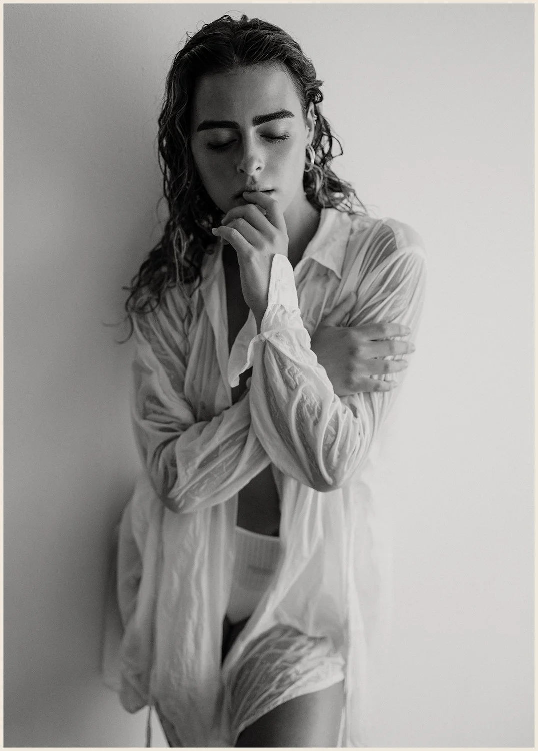 A black-and-white photograph of a young woman with curly hair, wearing a wet, partially open shirt and underwear, standing against a plain wall with her eyes closed, touching her lips with one hand and crossing her arms over her chest.