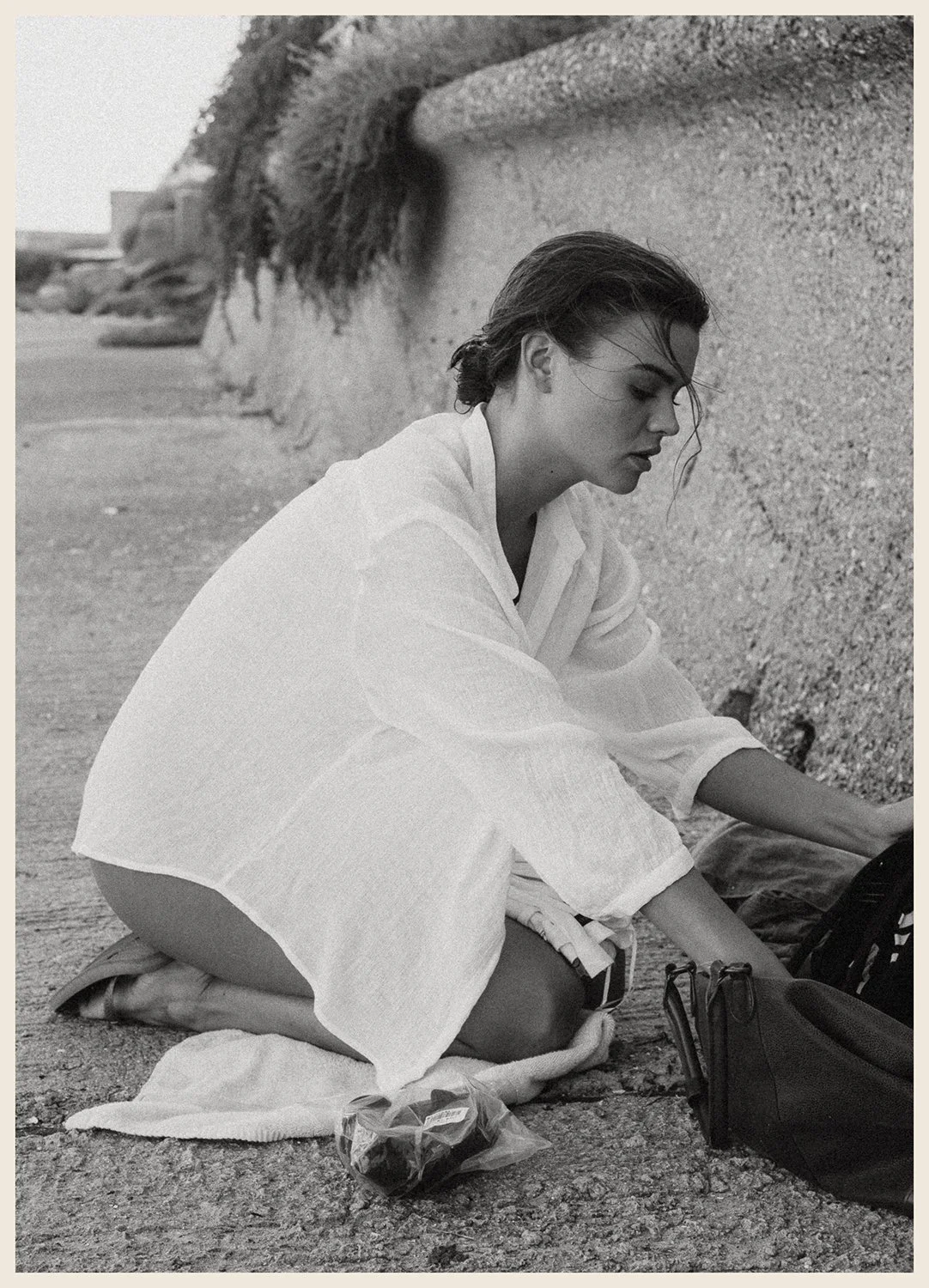 A woman kneeling on a beach in a white shirt, looking at her bag, with a pebble beach and rock wall in the background.