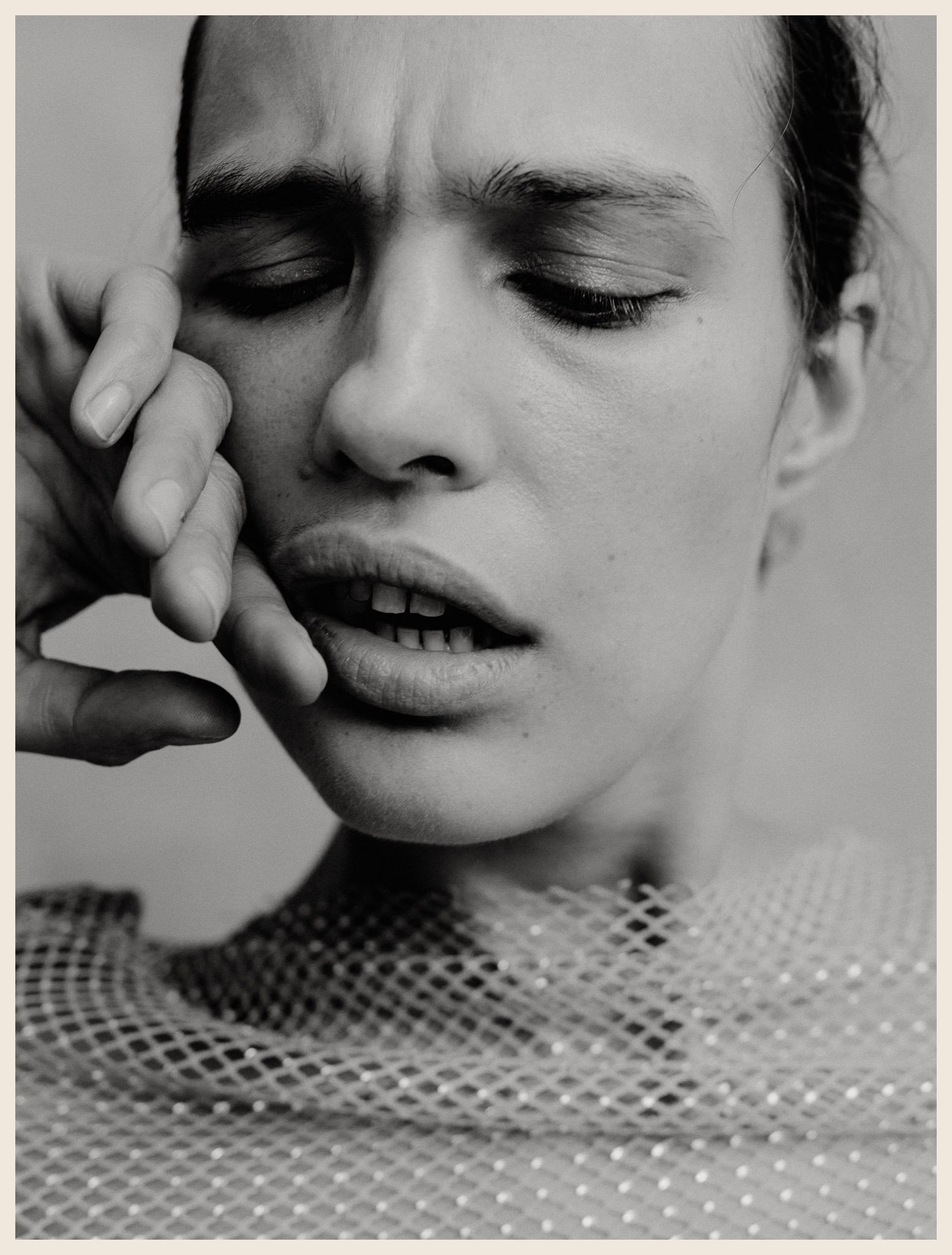 A black-and-white close-up photo of a woman with her eyes closed, mouth slightly open, and hand touching her face, wearing a sparkly mesh top.
