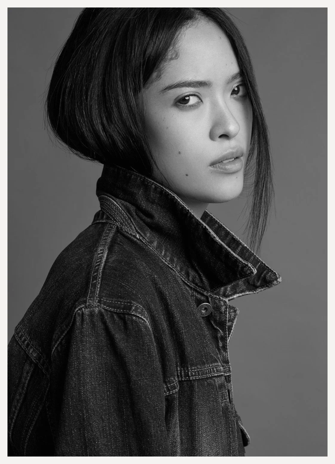 Black and white portrait of a young woman with dark hair wearing a denim jacket, looking at the camera.