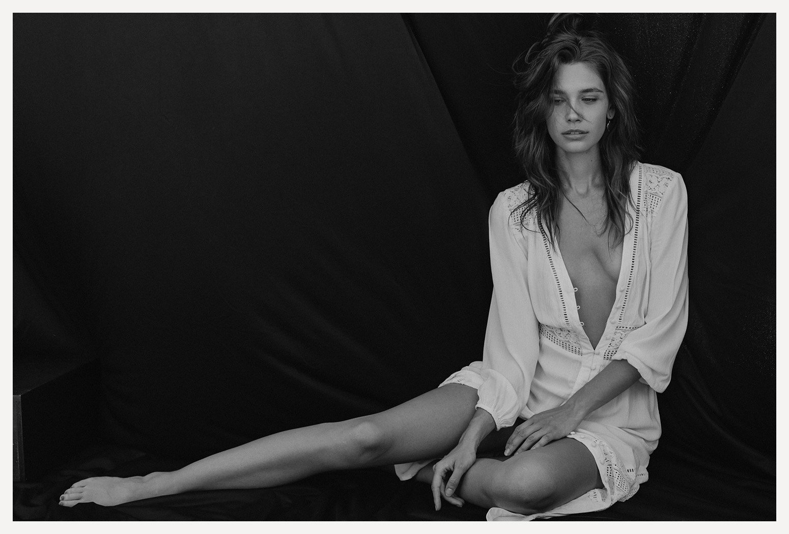 Black-and-white photo of a woman sitting on the floor against a dark backdrop. She is wearing a light-colored, loose dress with embroidery and lace details. Her hair is wavy and slightly tousled. She has a contemplative expression, looking down sligh