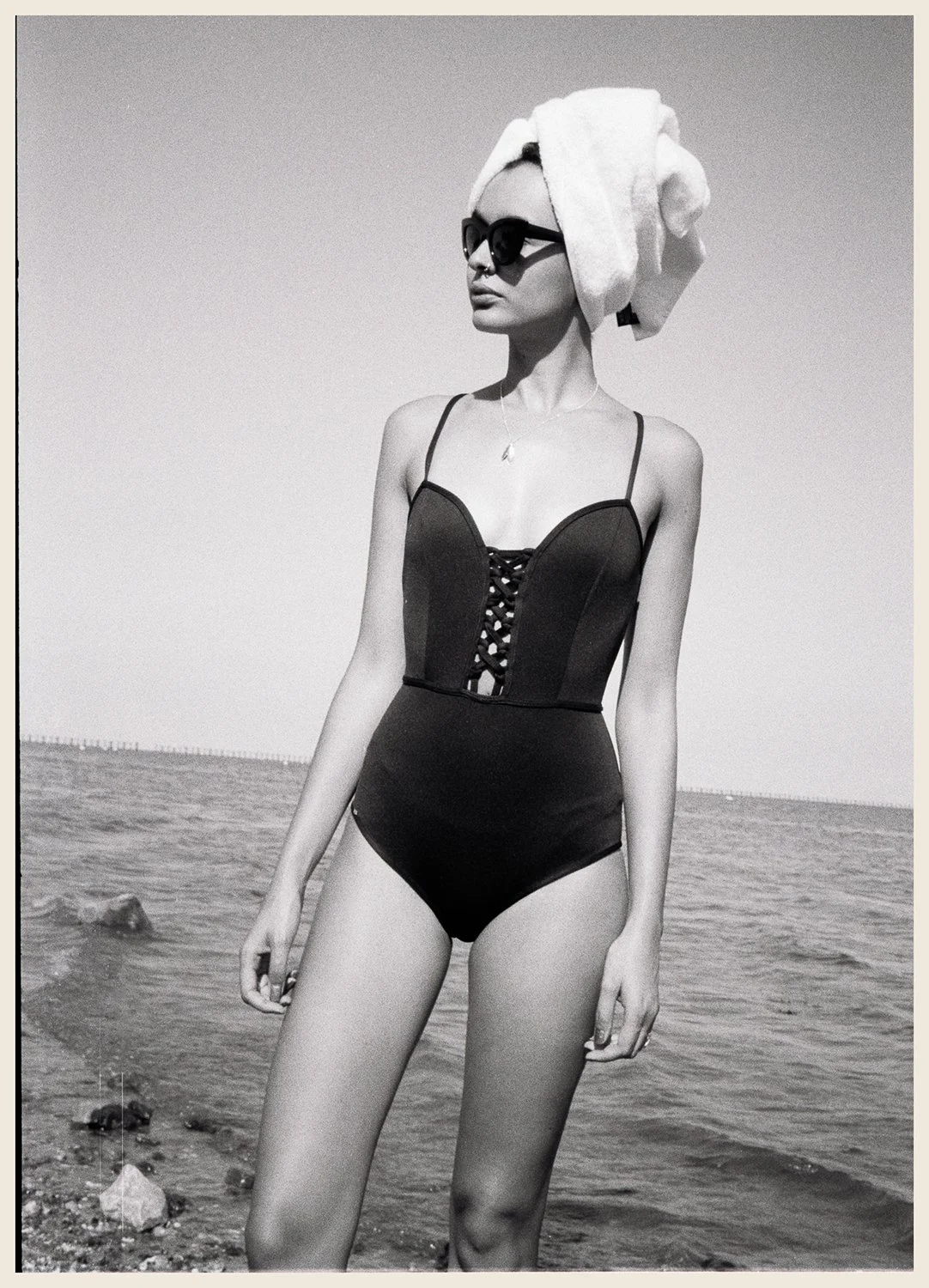A woman in a black swimsuit with lace-up front, wearing sunglasses and a towel wrapped around her head, standing on the beach with ocean and rocks in the background.