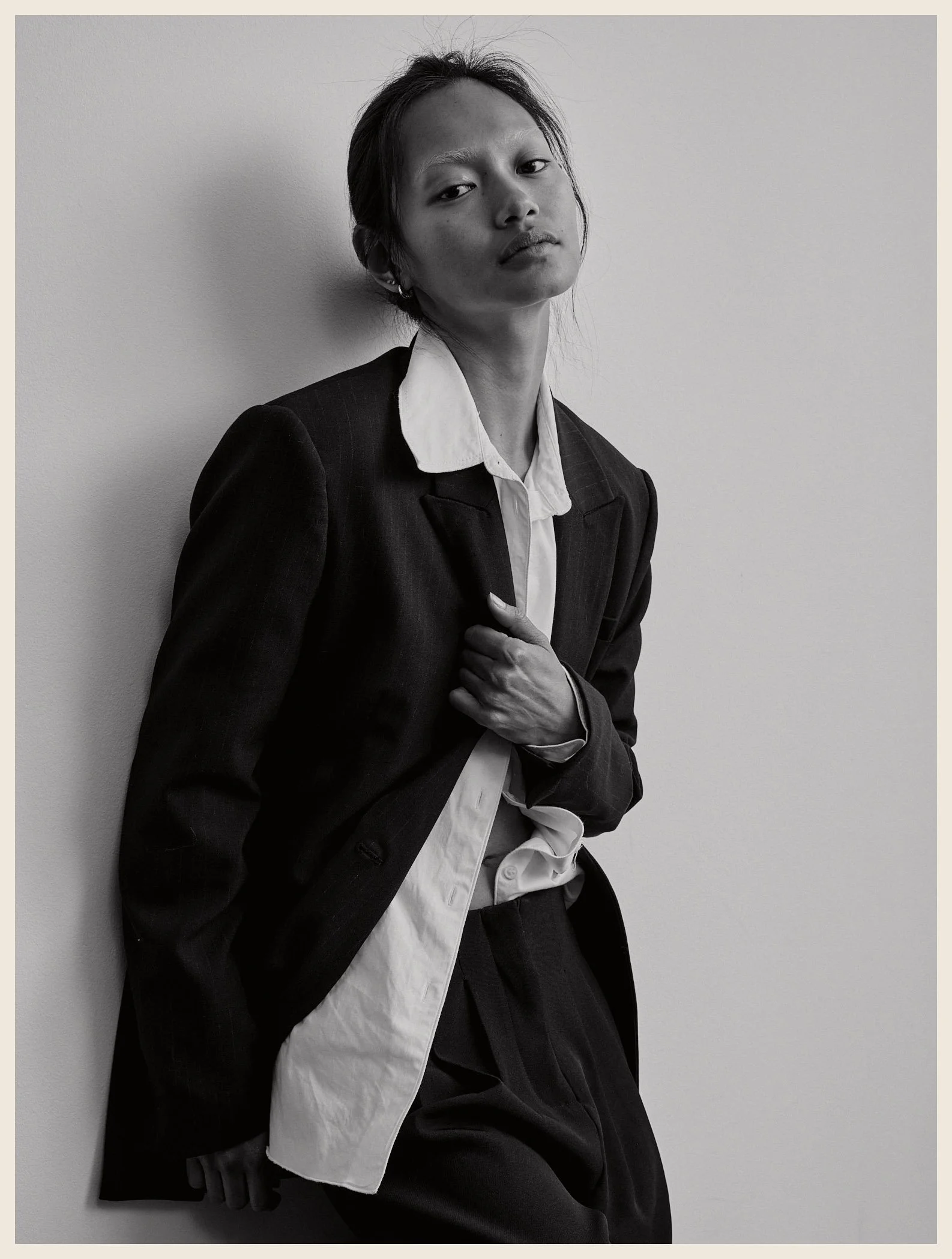 Black and white photo of a woman in a suit, leaning against a plain wall, with her hand on her chest and a serious expression.