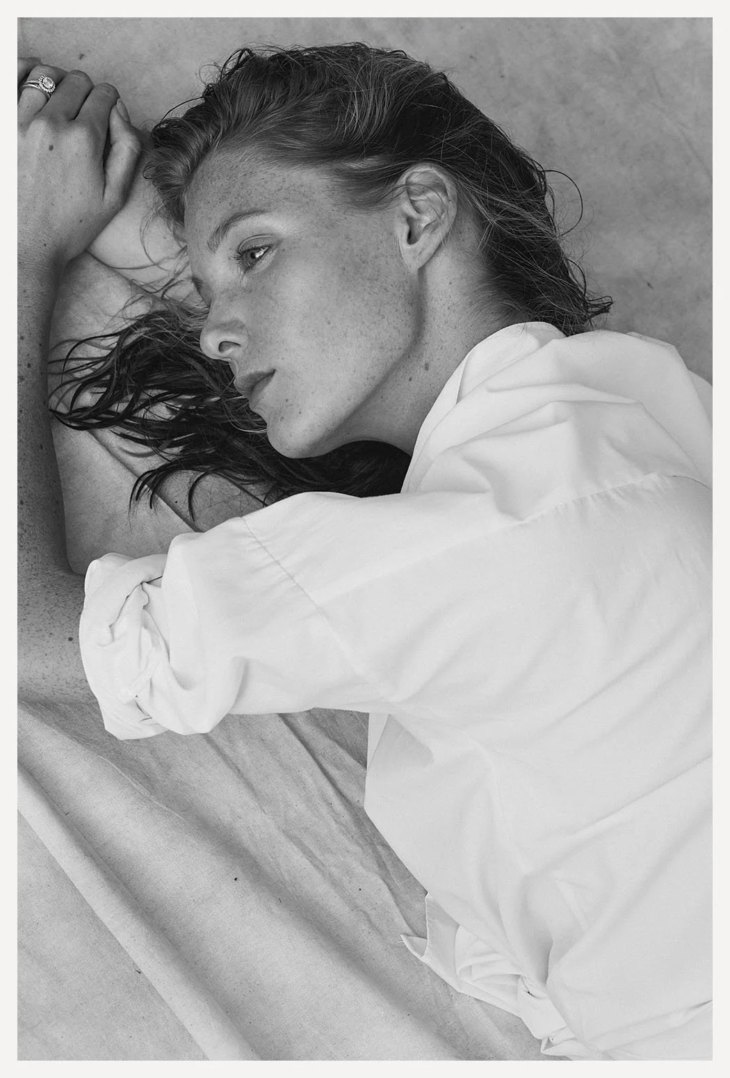 Black and white photo of a woman with freckled skin lying down with her eyes closed, resting her head on her arm, wearing a white shirt.