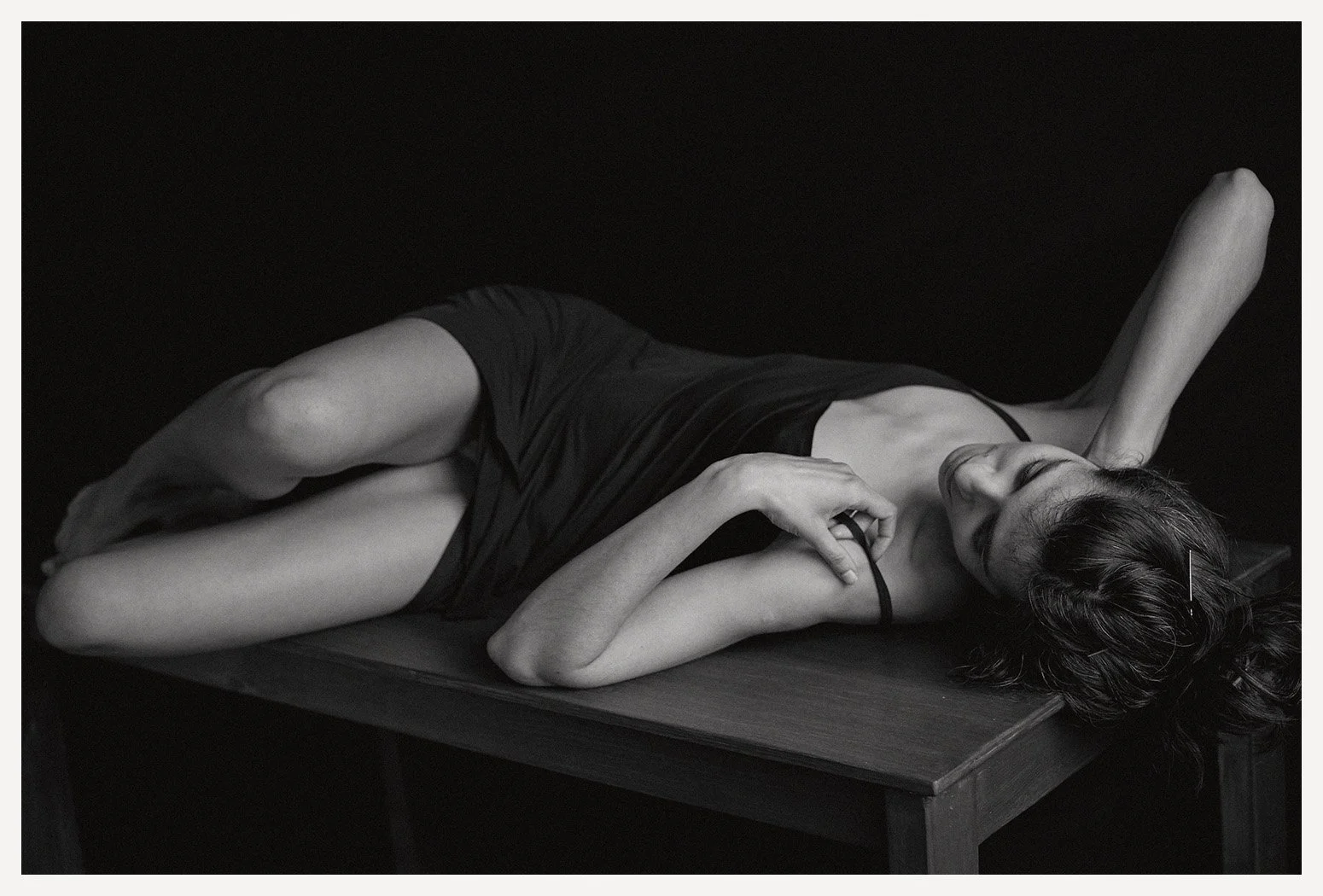 A black and white photograph of a woman lying on a wooden table, wearing a black dress, with her eyes closed and one hand resting on her chest.