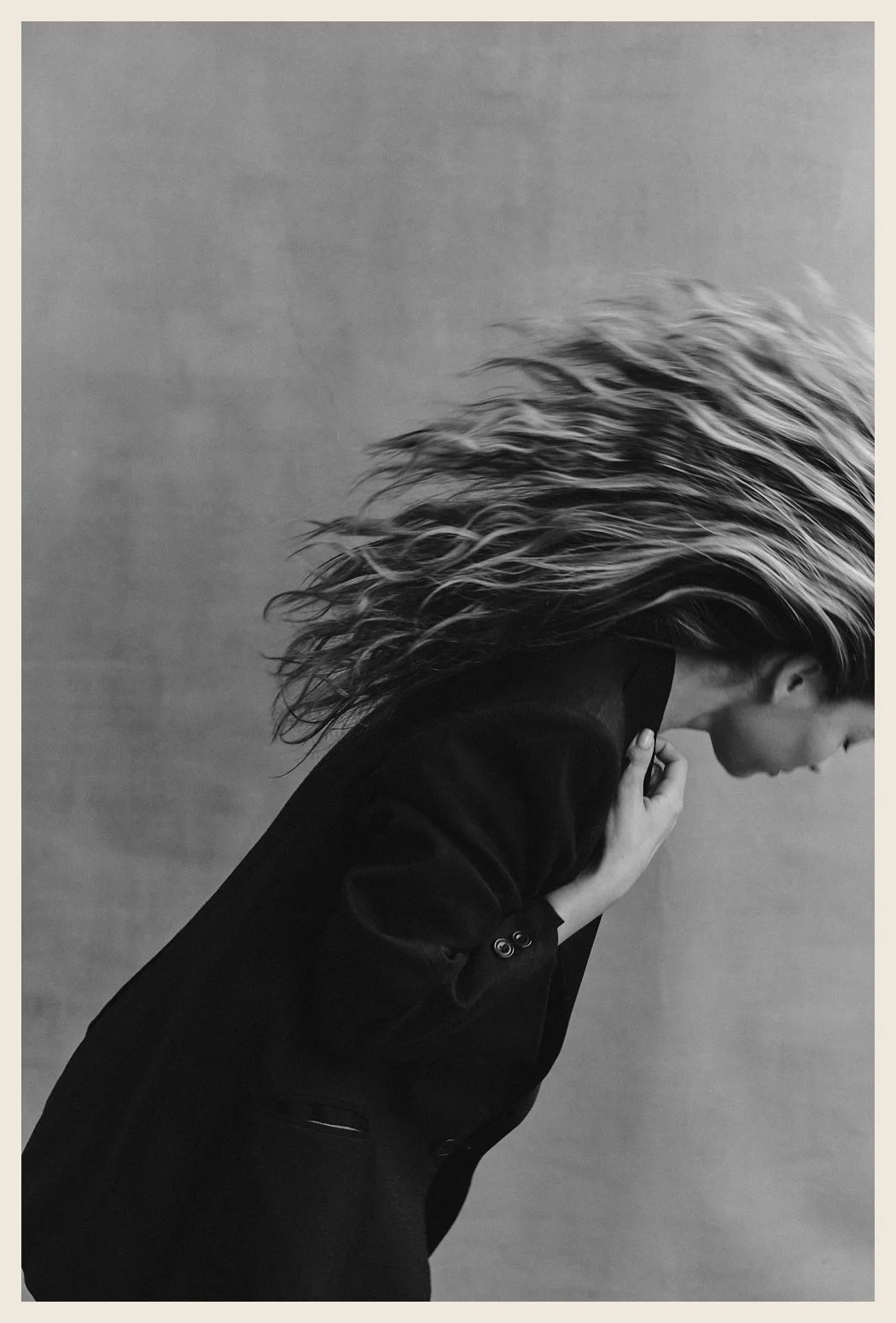 Black and white photo of a woman with long, wavy hair, wearing a dark blazer, leaning forward with her hand near her face, against a plain background.