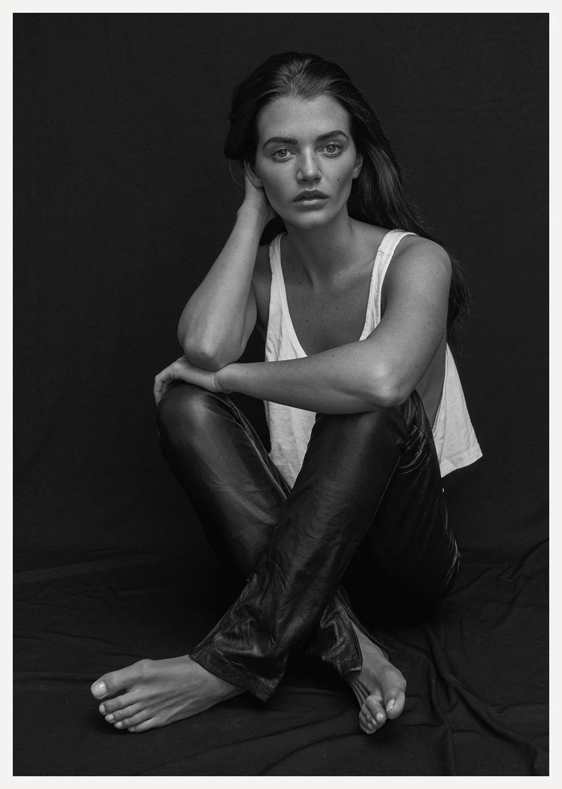 Black and white photo of a woman sitting on a dark fabric surface, dressed in a sleeveless top and leather pants. She has long hair and is looking directly at the camera with a contemplative expression, one hand resting on her neck and the other on h