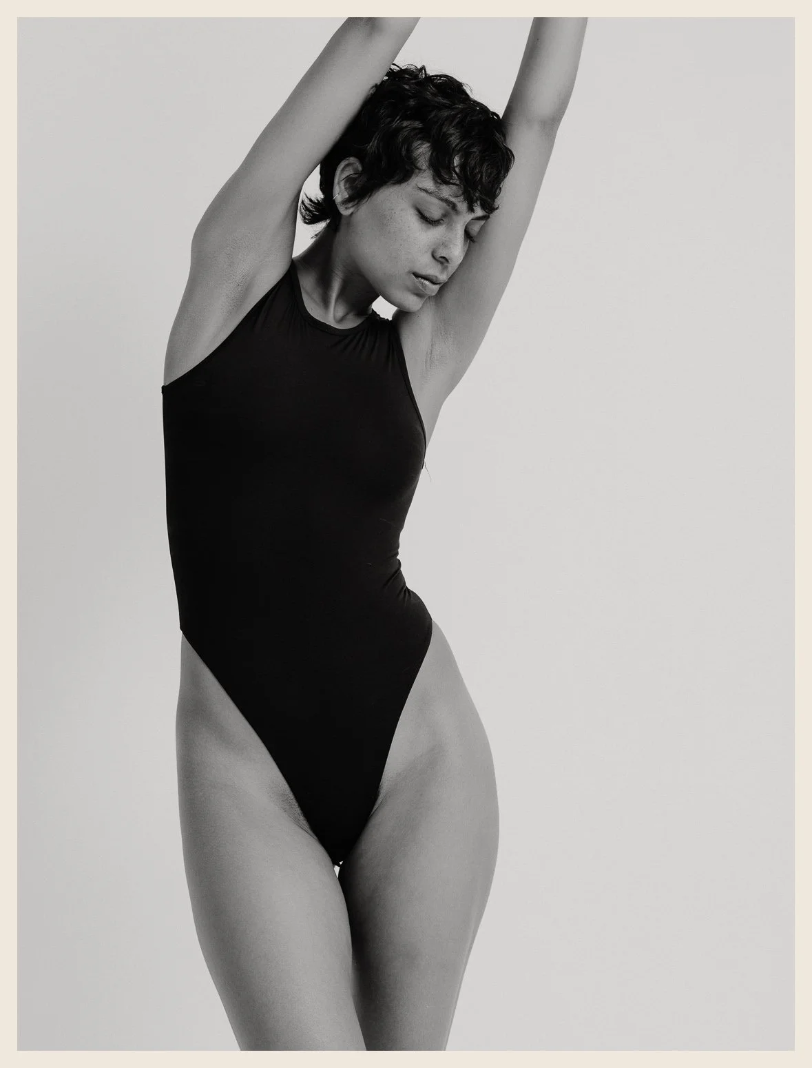 A woman in a black swimsuit with her arms raised overhead, looking down, against a plain light background.