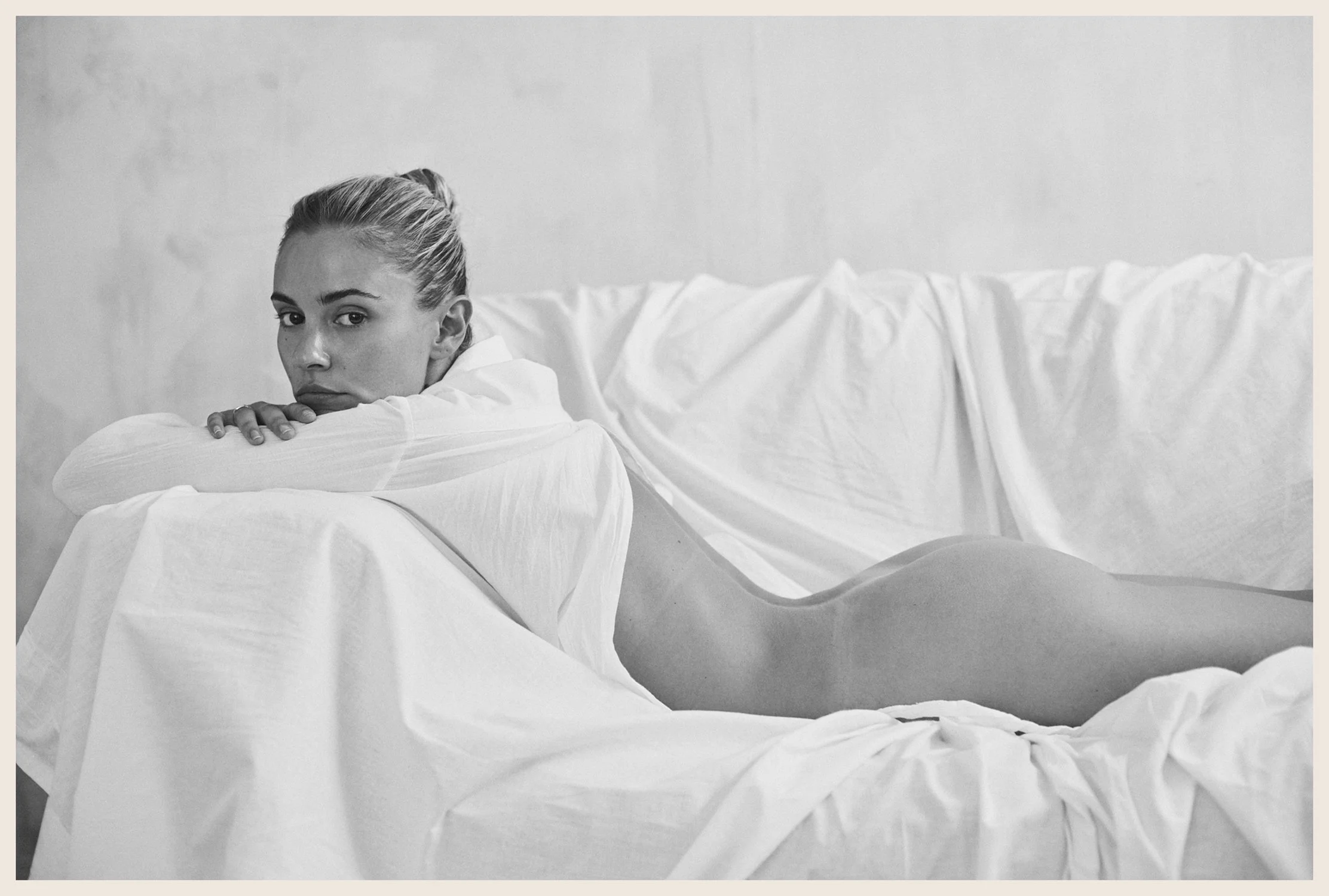 A black-and-white photograph of a woman lying on her stomach on a bed with white sheets, resting her head and arms on the bed, and looking at the camera.