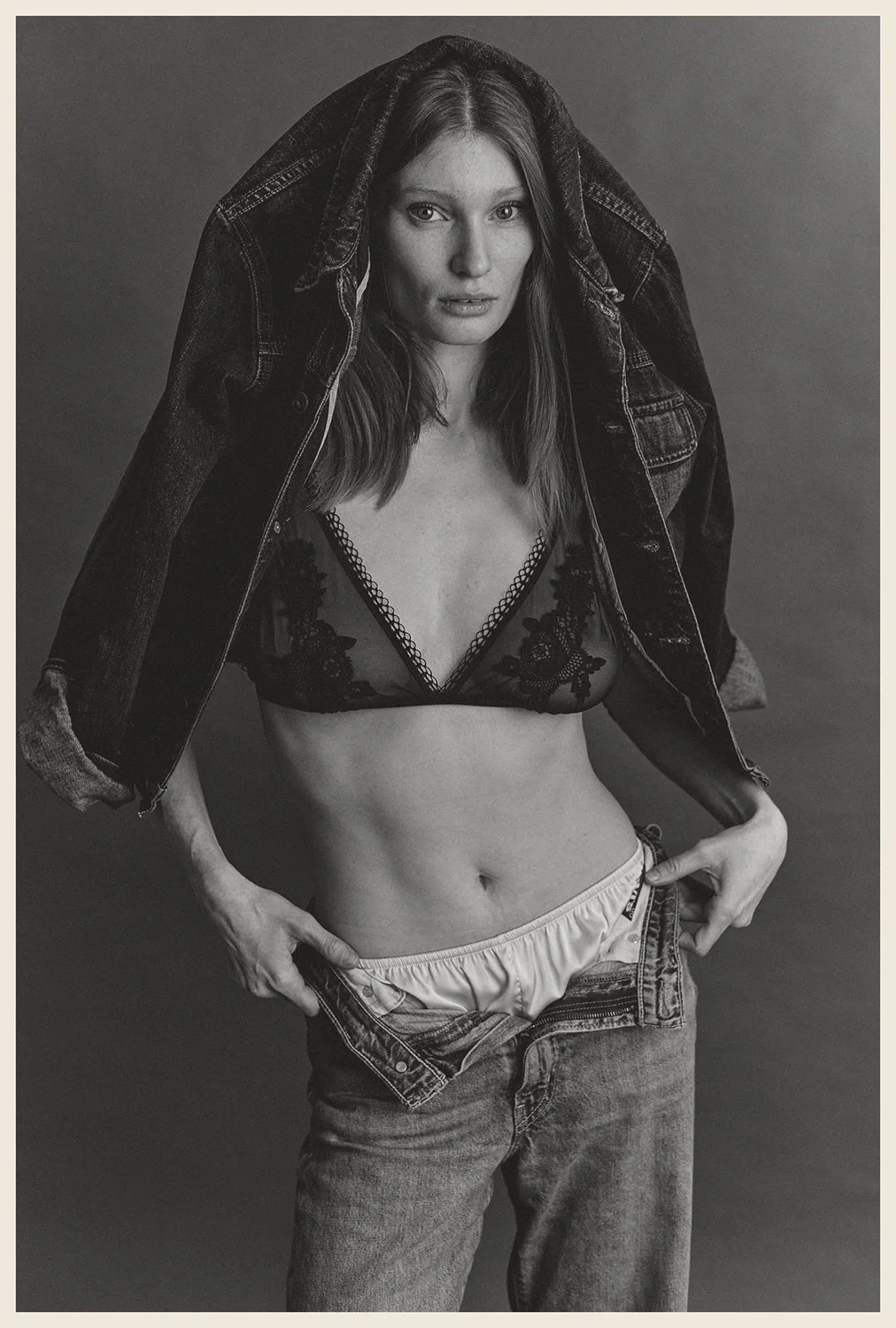 Black and white portrait of a woman with long hair, wearing a lace bralette, partially unbuttoned jeans, and a denim jacket draped over her shoulders, against a plain background.