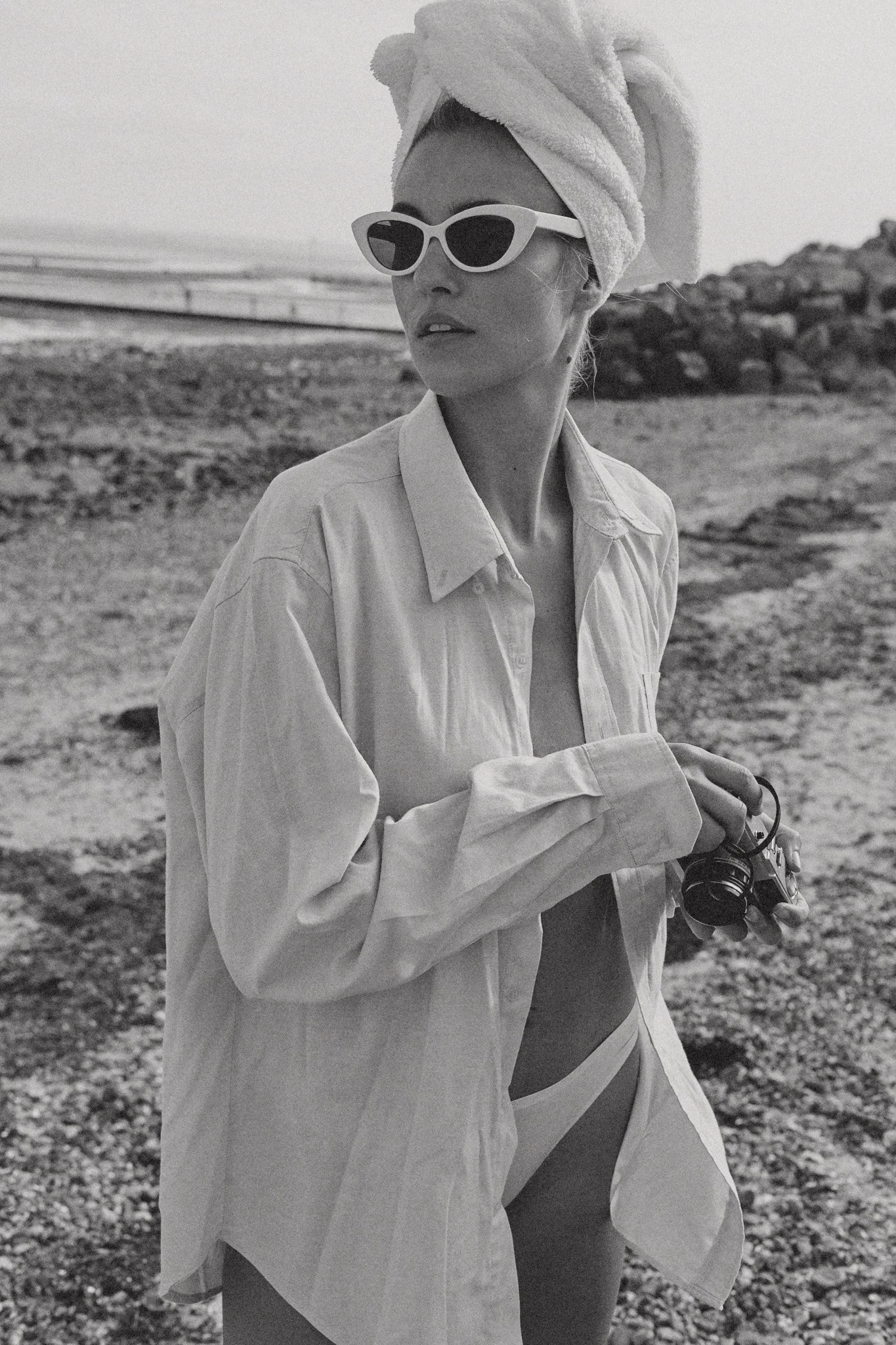 Woman on a rocky beach wearing sunglasses, a towel wrapped around her head, an oversized shirt, and holding a camera.