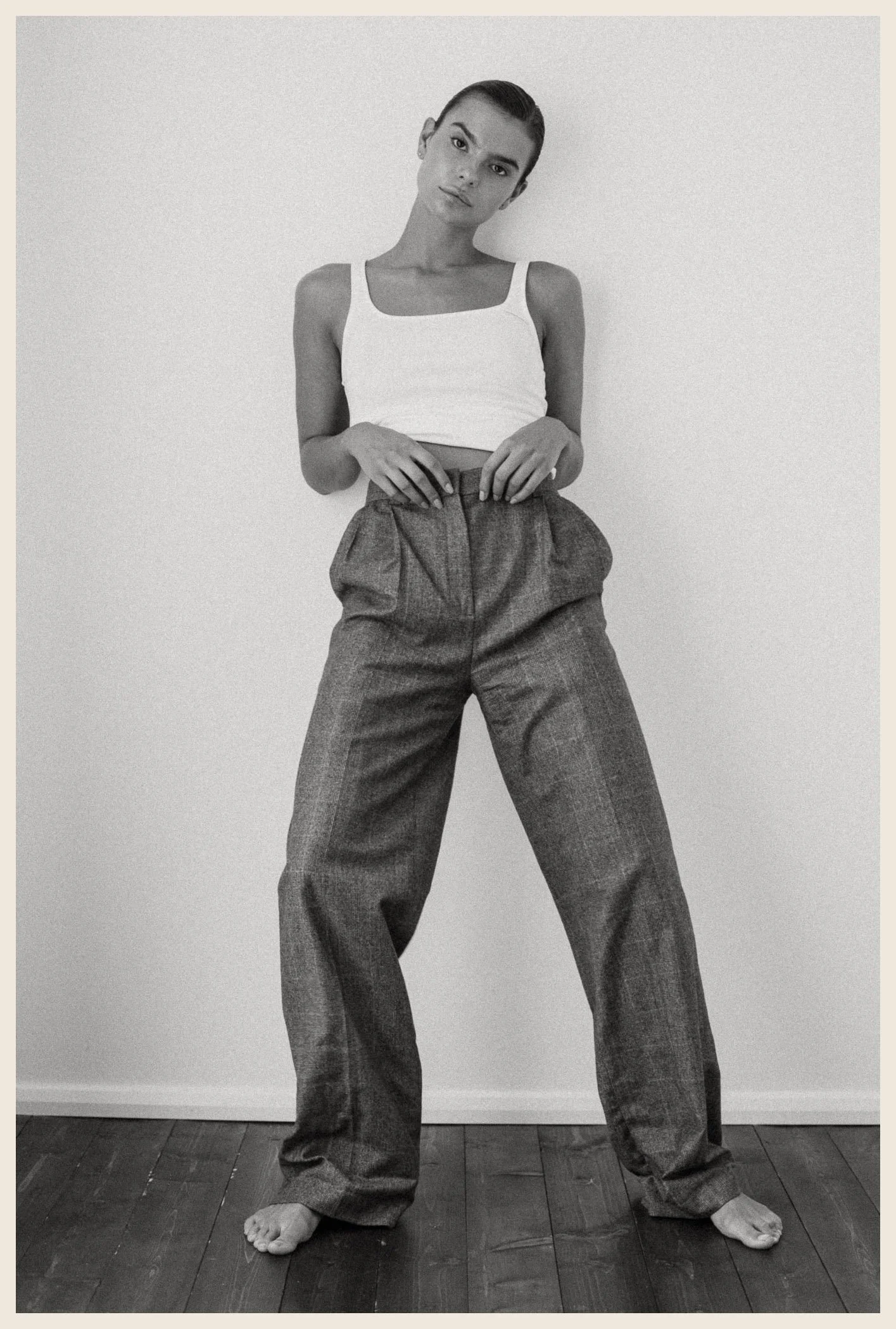A woman standing against a plain wall, wearing a white tank top and loose trousers, with her hair in a slicked-back style, looking at the camera.