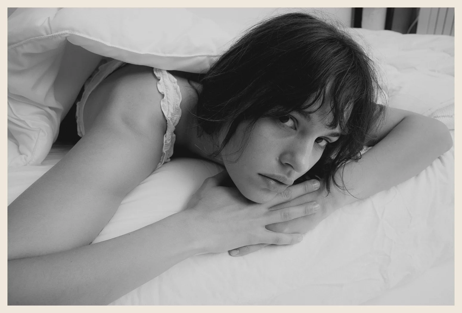 Black and white photo of a woman lying on a bed, resting her head on her arm and gazing at the camera.