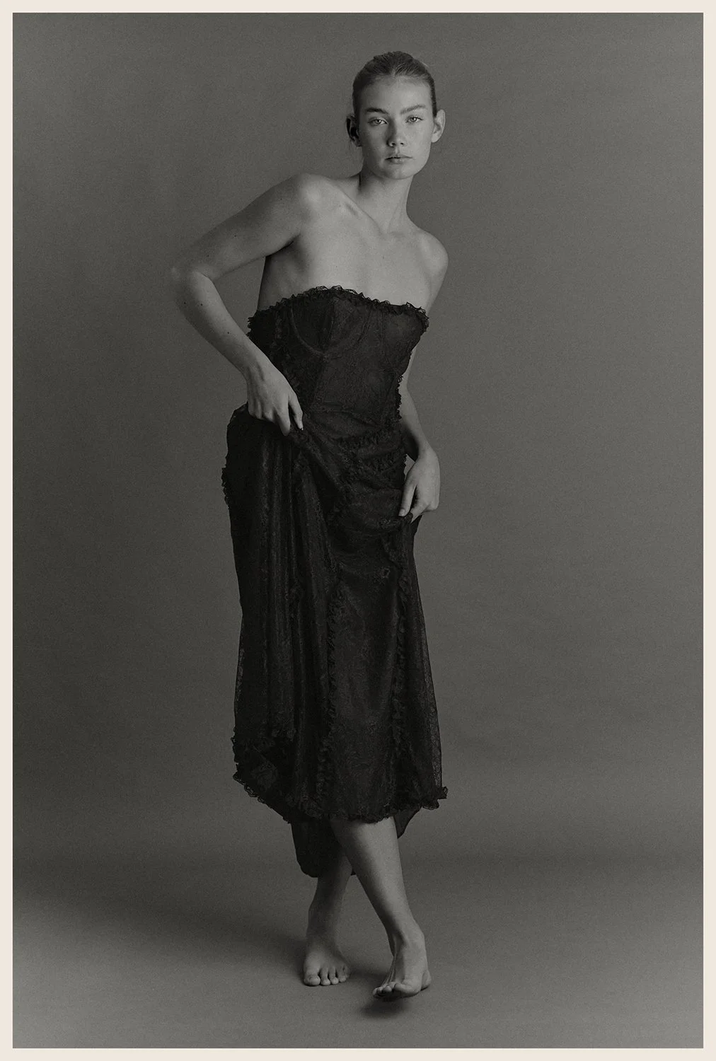 A black and white photo of a woman standing against a plain background, wearing a strapless black lace dress, holding the dress with one hand, and standing barefoot with one foot crossed over the other.