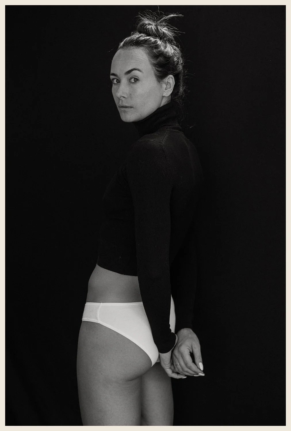 A black and white portrait of a woman with her hair in a messy bun, wearing a black turtleneck and white underwear, posing against a dark background.