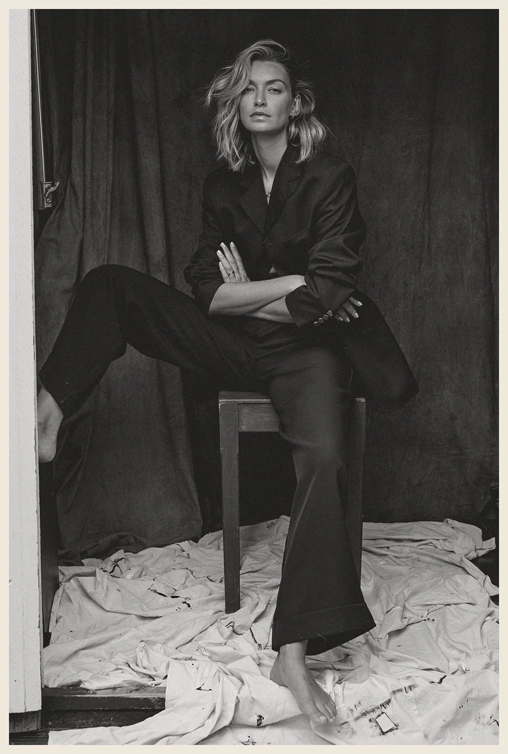 A black-and-white photo of a woman sitting on a chair with one leg across the other, wearing a blazer and wide-leg pants, in front of a dark curtain, with crumpled paper on the ground.