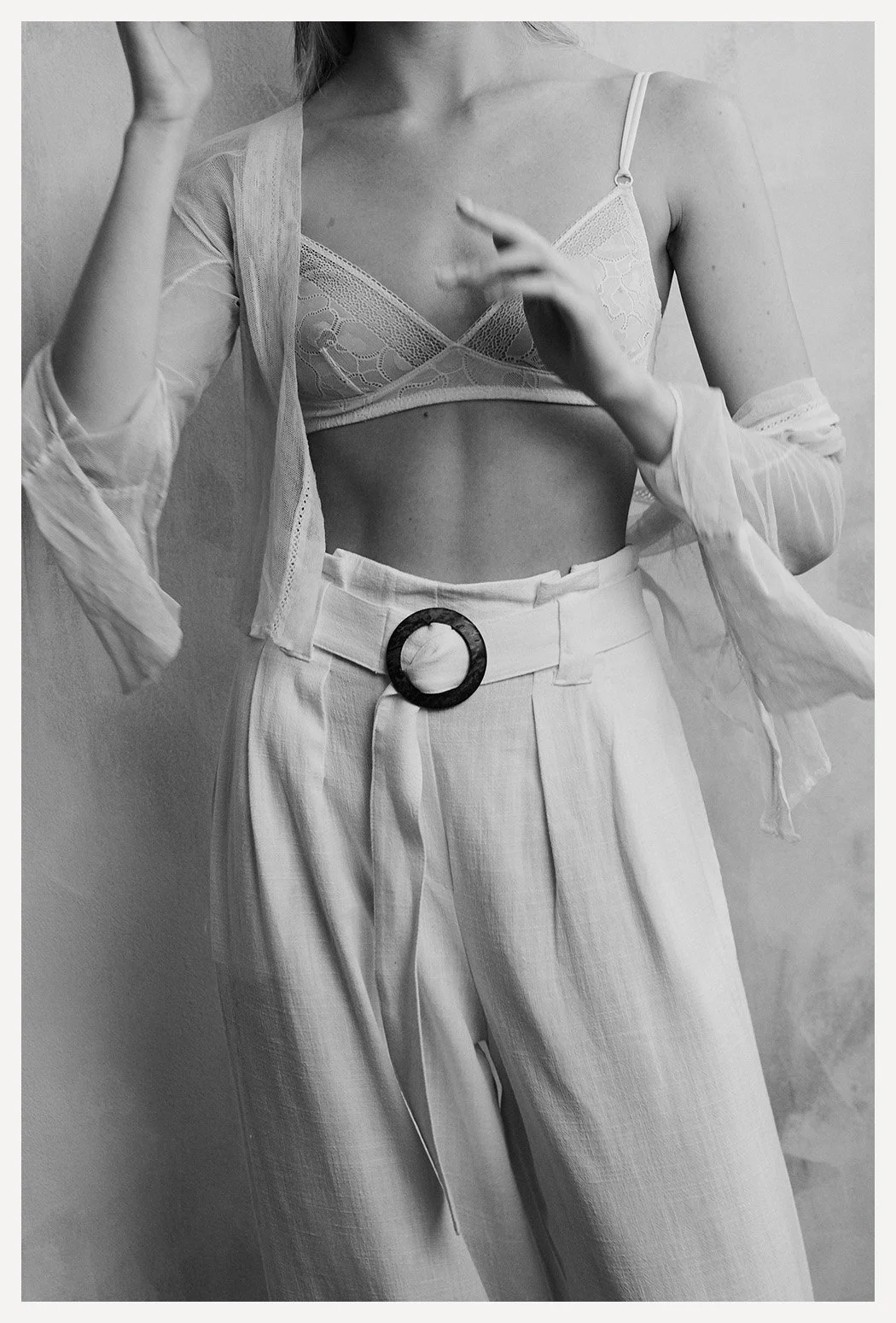 Black and white photo of a woman wearing high-waisted pants with a belt, a lace bralette, and a sheer jacket, with her hands partially in frame.