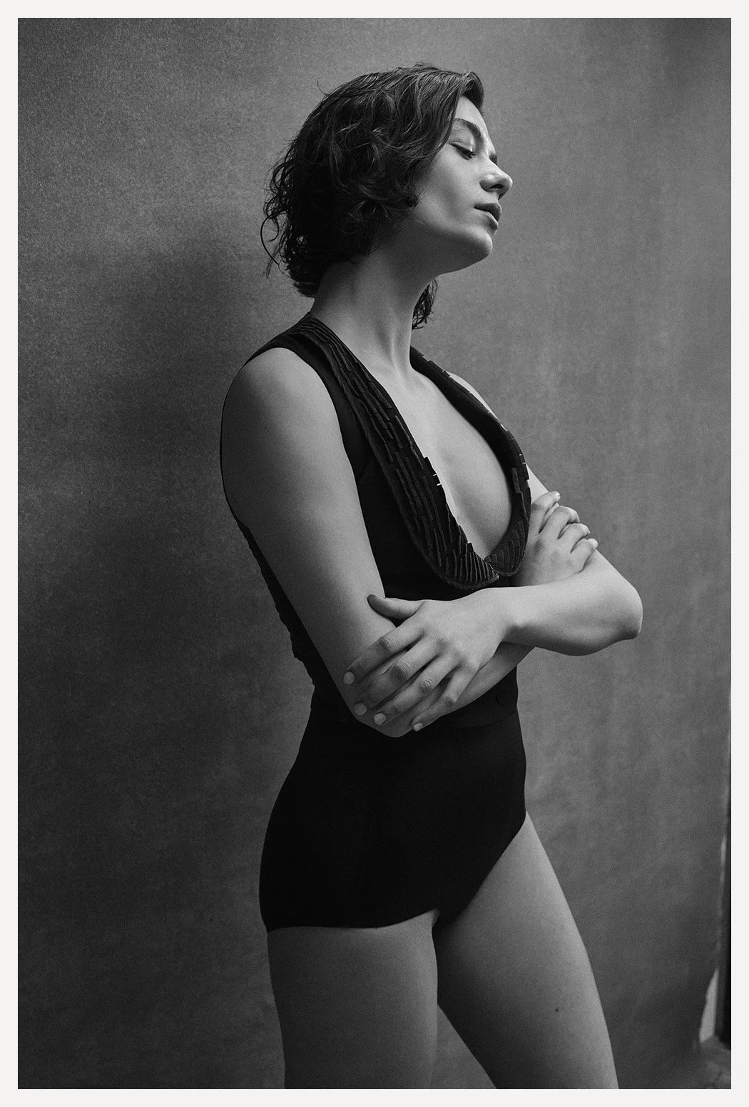 A black and white portrait of a woman with short wavy hair, wearing a sleeveless top and high-waisted pants, standing with arms crossed and eyes closed against a plain background.