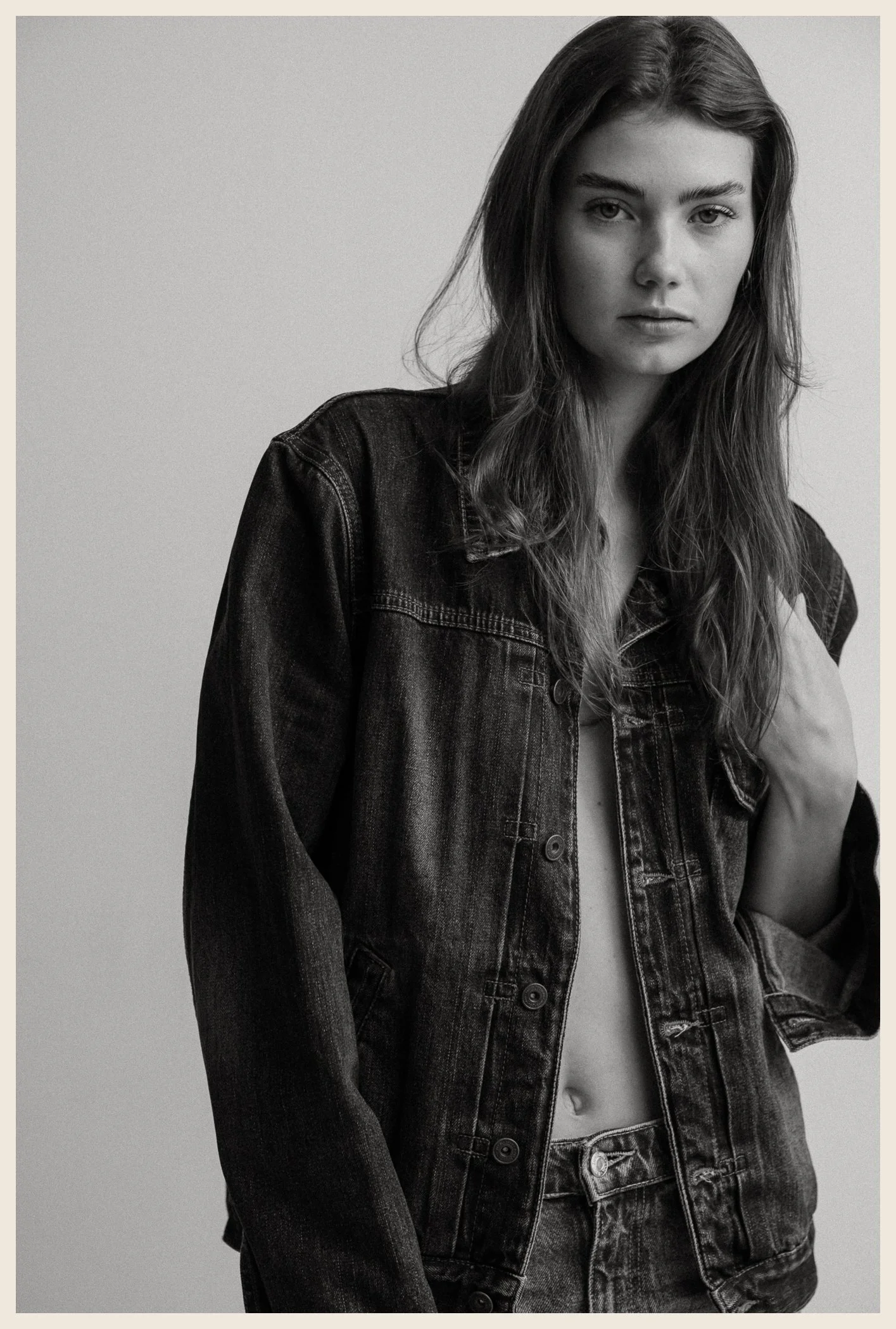 Black-and-white photo of a young woman with long hair, wearing an unbuttoned denim jacket. She is looking directly at the camera with a serious expression.