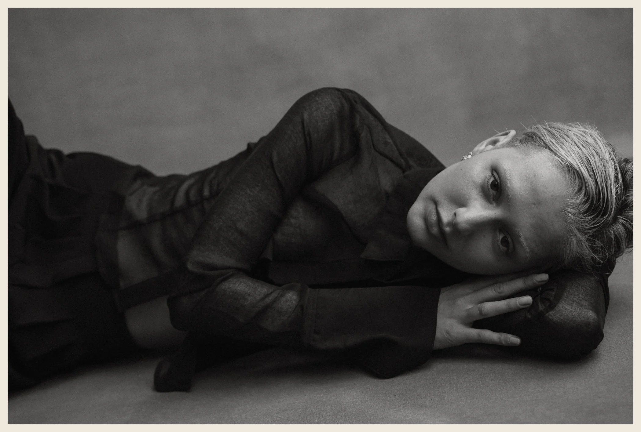 A black and white photograph of a woman lying on her side on a flat surface, resting her head on her arm, looking at the camera. She has short, styled hair and is wearing a sheer, long-sleeved top.