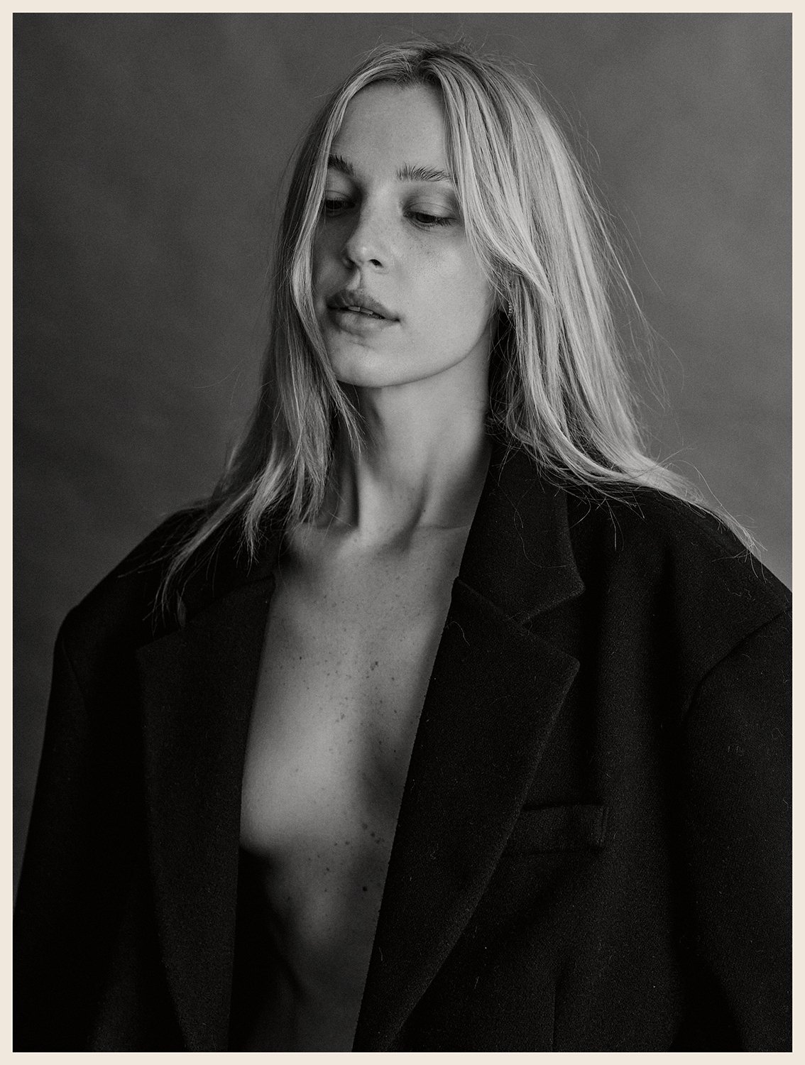A black-and-white portrait of a young woman with light hair, wearing an open blazer, looking down and away against a plain background.