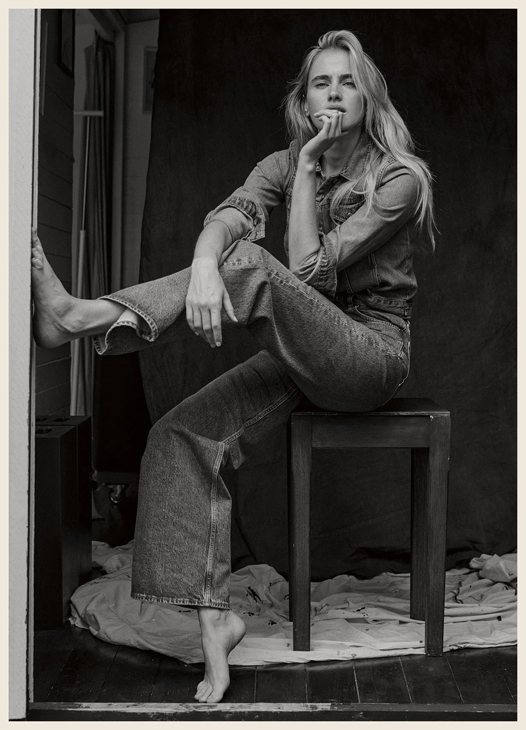A black and white photo of a young woman sitting on a wooden stool with one leg crossed over the other. She is barefoot, wearing denim jeans and a denim shirt. She has long, wavy hair and a serious expression, with her chin resting on her hand. The b