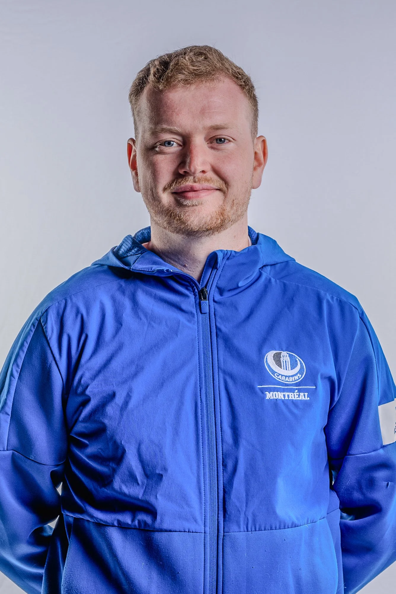 Portrait of a man with light skin, short light brown hair, and a beard, wearing a blue sports jacket with 'Montreal' and a logo on the chest, posing against a plain light background.