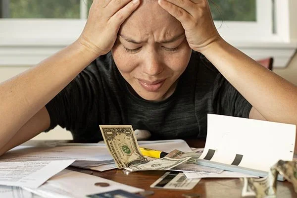 woman head in her hands face looking scared with cash and credit cards and bills piled on the table in front of her