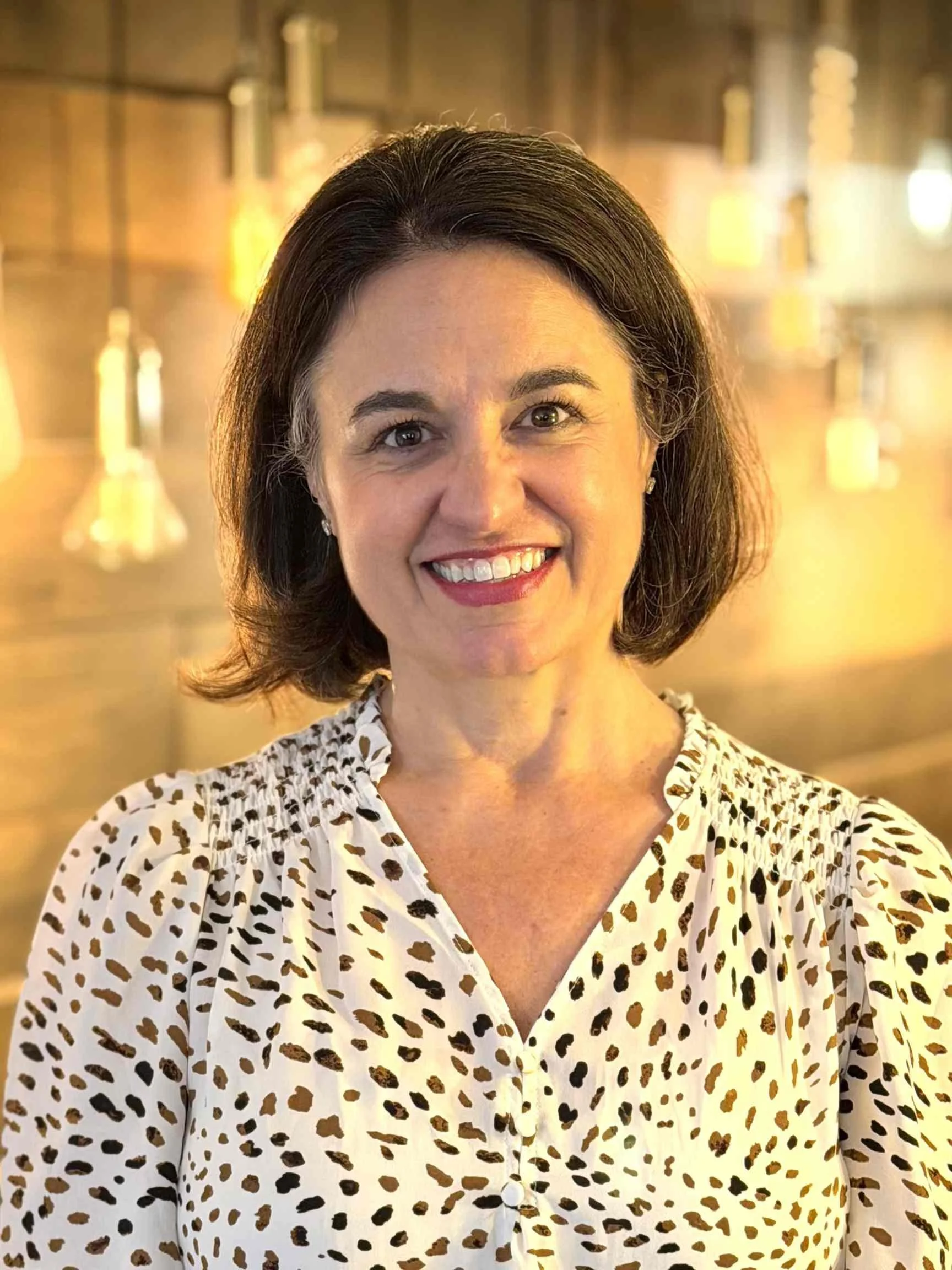 A woman with shoulder-length dark hair, glasses, red lipstick, wearing a denim shirt, smiling, with warm lighting and wooden background fixtures