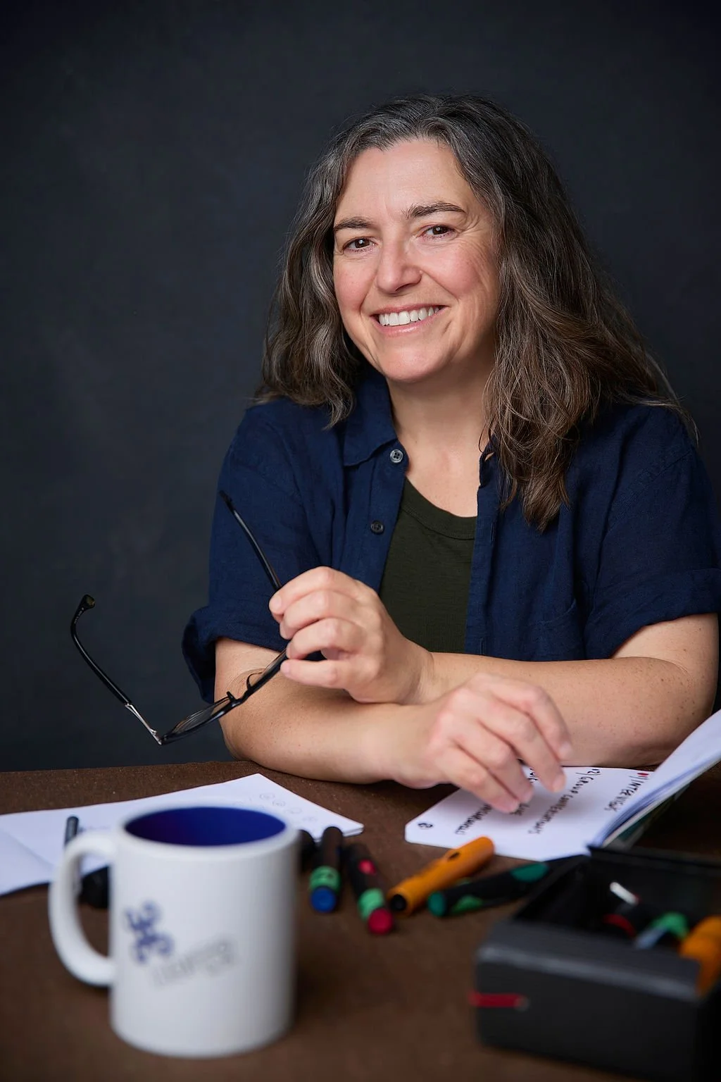 A smiling white woman with brown hair streaked with white, holding glasses in one hand, leaning on a table.