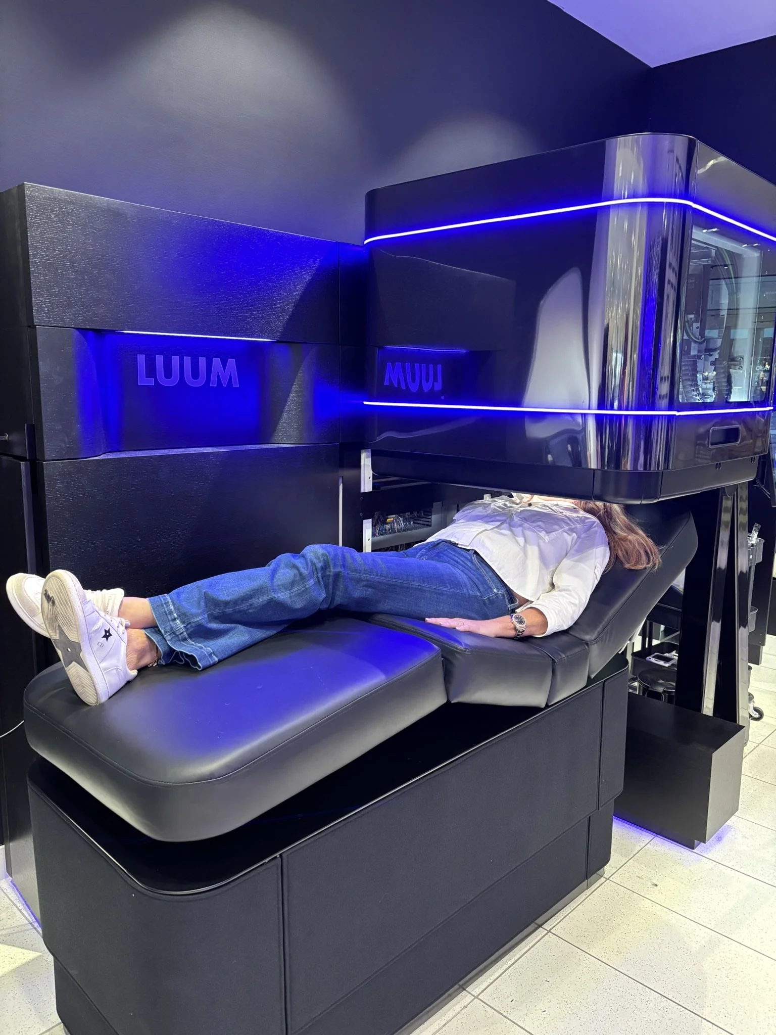 Person lying on a cushioned lounge chair inside a transparent capsule structure in a shopping mall, with decorative pillows and a stool nearby.