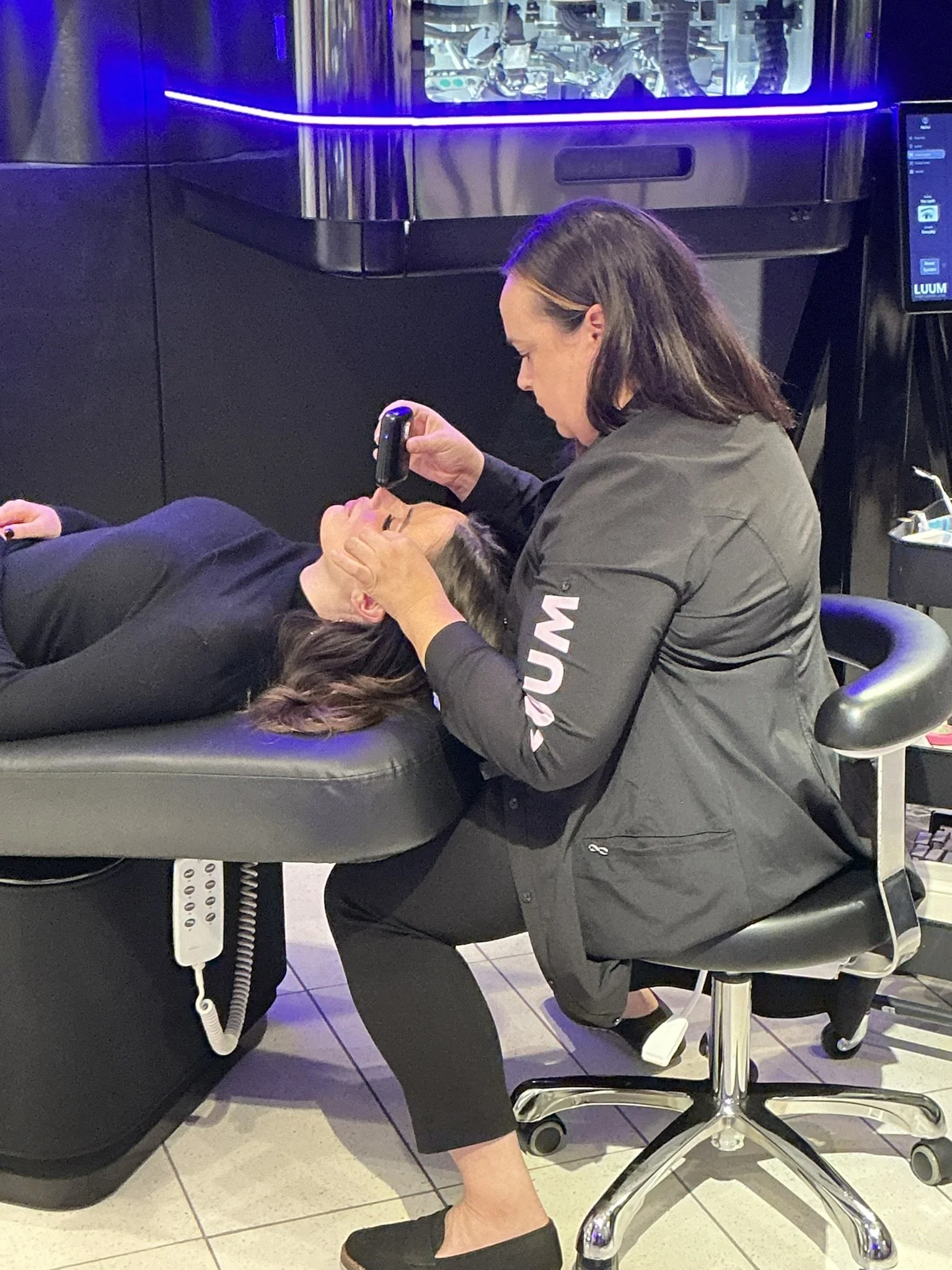 A woman lying on a medical examination table with her head to the side, while a healthcare professional examines her forehead with a device in a modern medical facility.