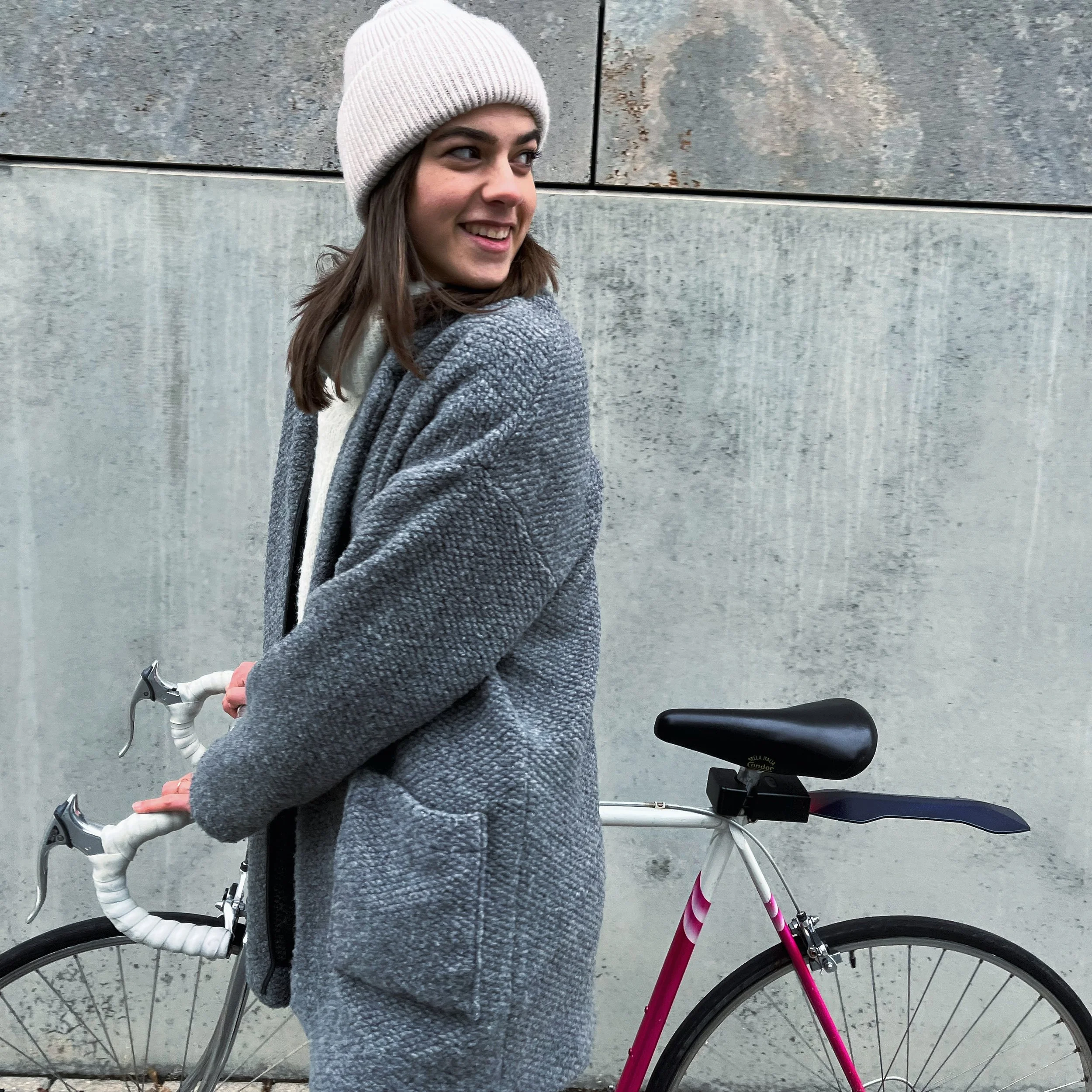 Person in gray coat and white beanie with bicycle, outside concrete wall, smiling.