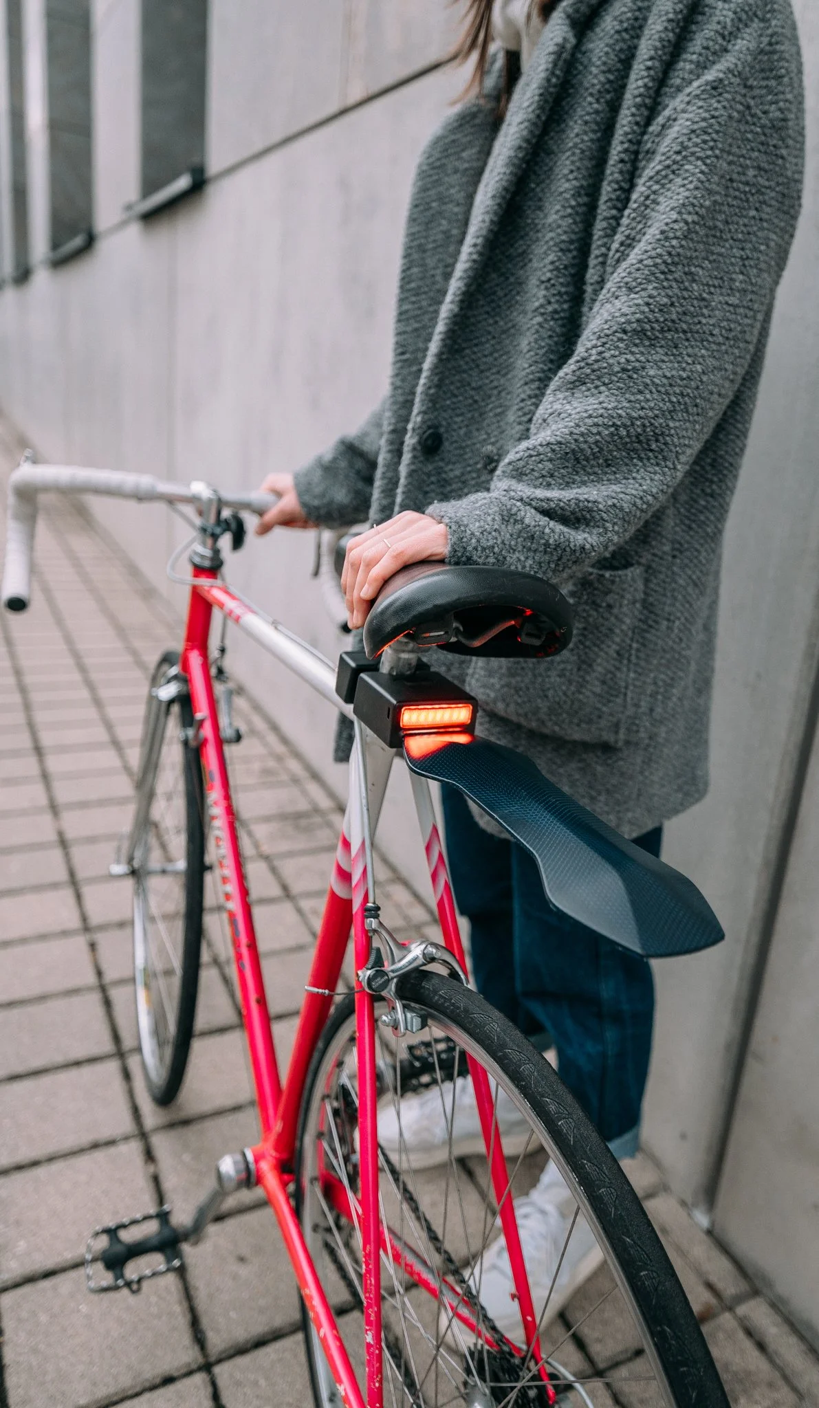 Person in a gray coat holding a red bicycle with a mudguard and rear light, standing on a tiled pathway.