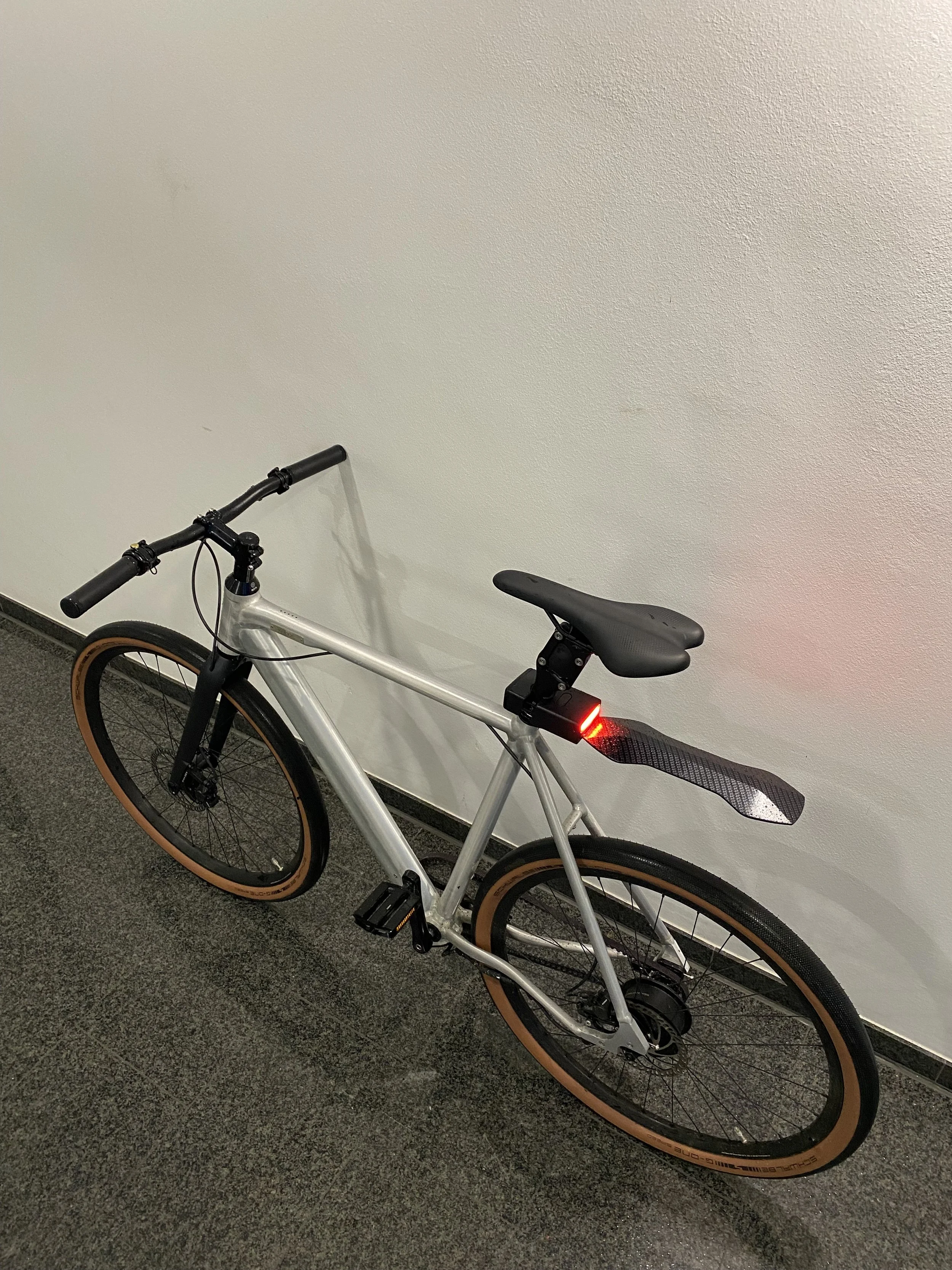 A silver bicycle with black handlebars and tan sidewall tires, parked indoors on a tiled floor. It features a rear fender and a red rear light mounted below the seat.