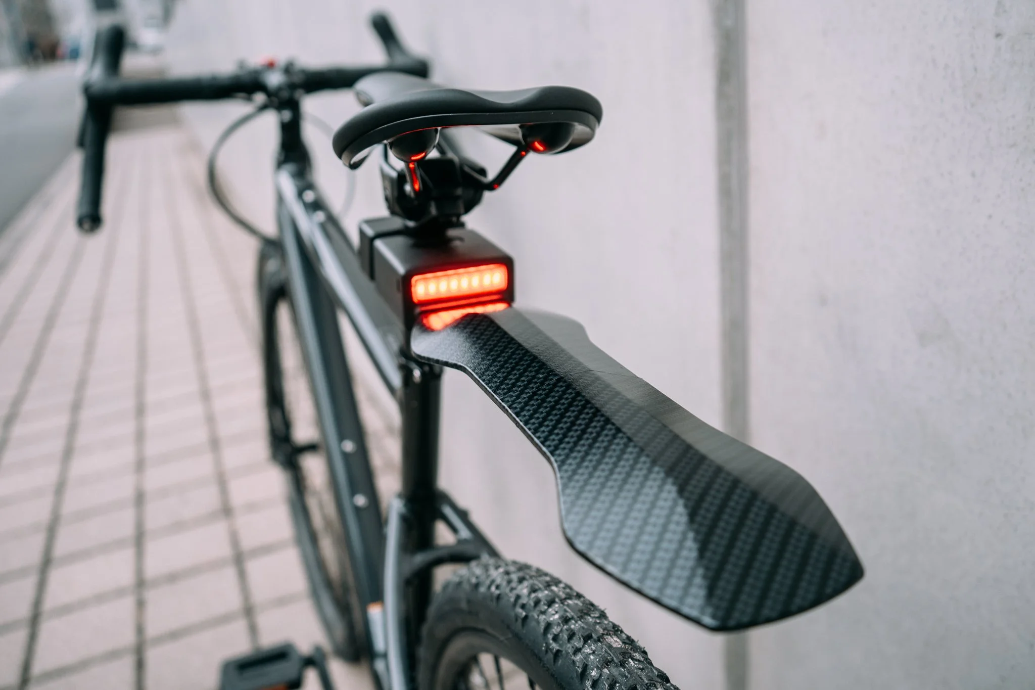 Close-up of a black bicycle's rear section, featuring a red LED taillight and a carbon fiber fender.
