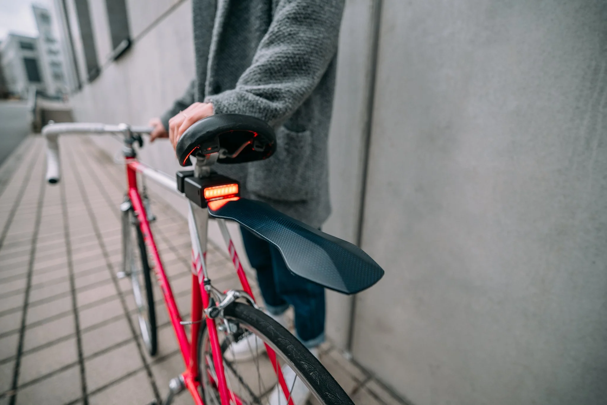 Person holding a red and white bicycle with a rear mudguard and an illuminated rear light next to a concrete wall.