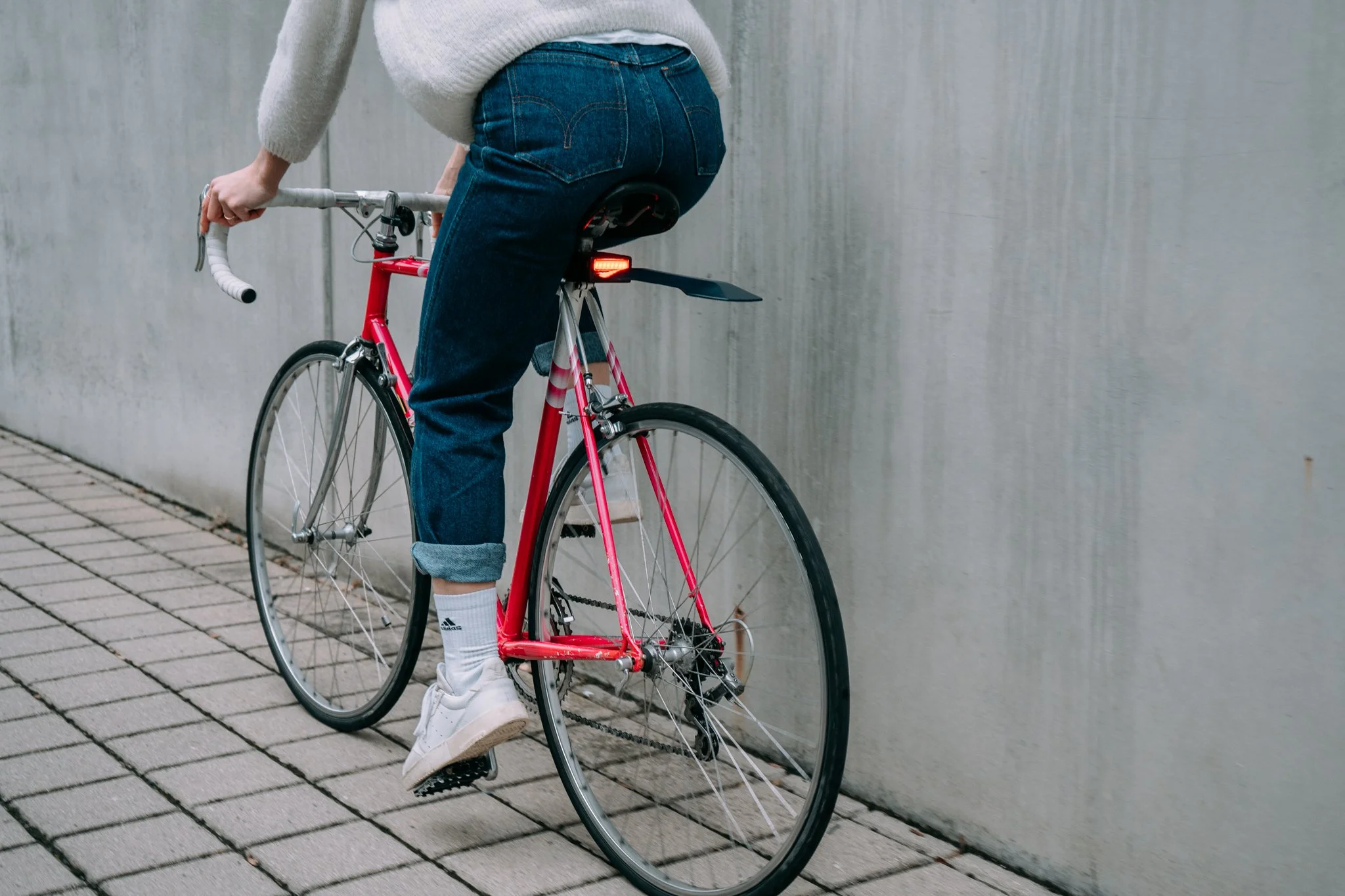 Person riding a red road bike with jeans and white sneakers on a paved path next to a gray wall.