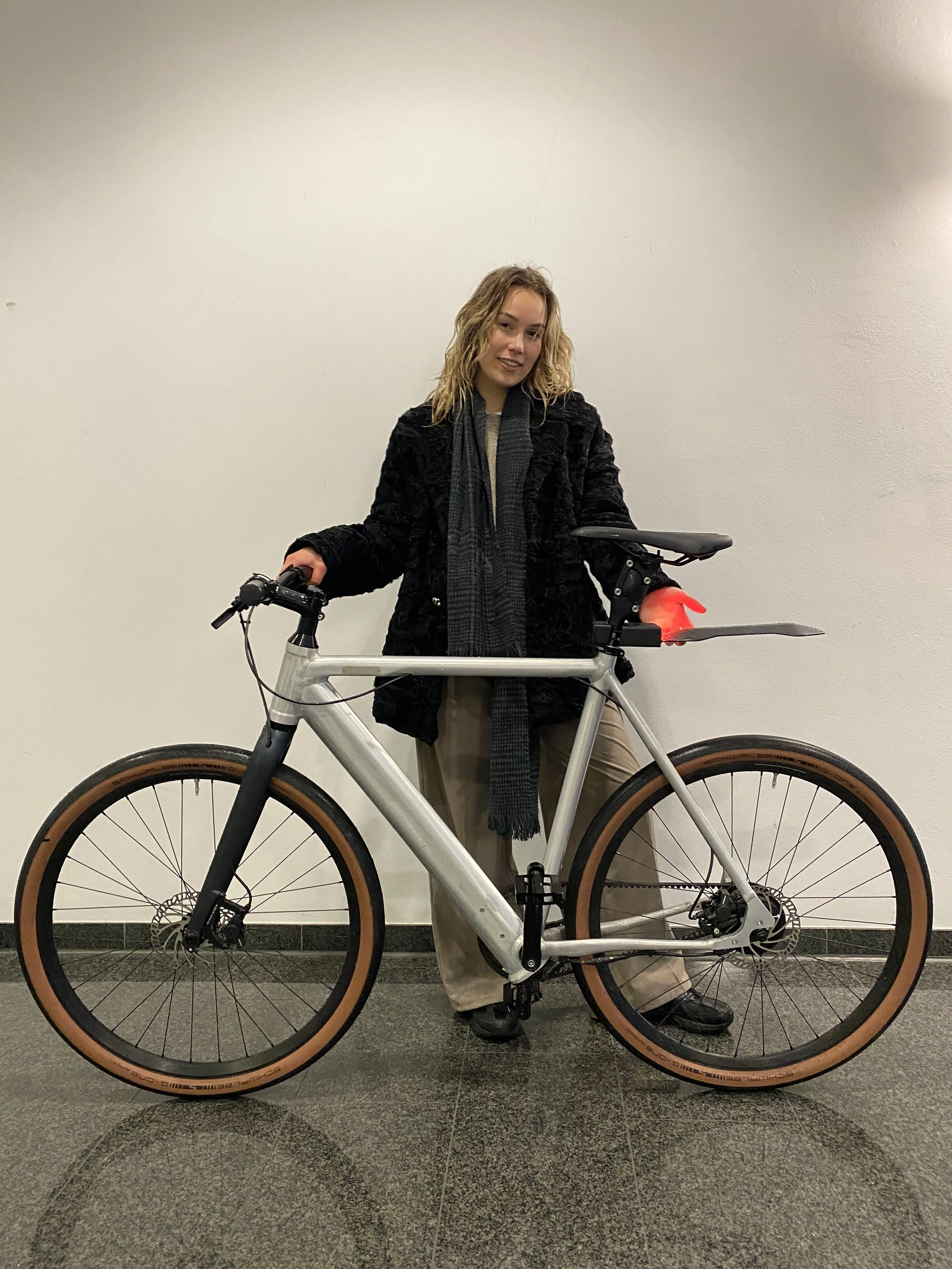 Person wearing a black coat and holding a bicycle with brown tires, standing indoors against a plain wall.