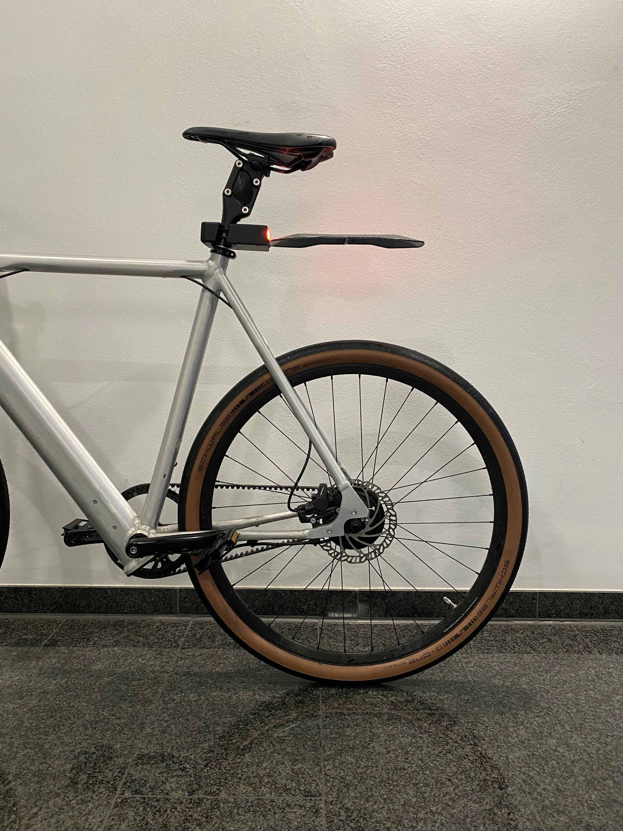 Rear view of a modern bicycle showing the back wheel, seat, and part of the frame. The bike features a rear light and a mudguard. The wheel has a tan sidewall tire and disc brake.