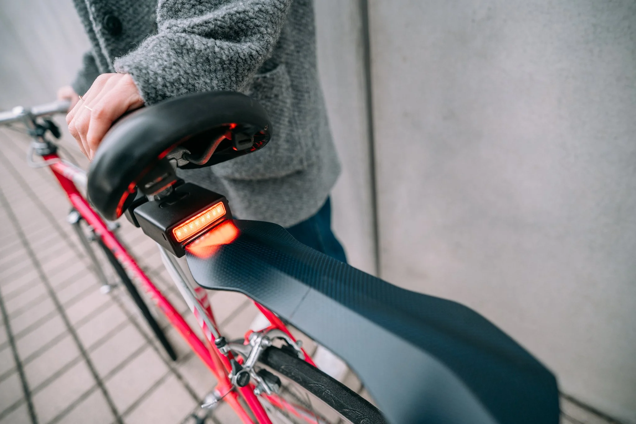 Person holding a red bicycle with a rear light on, wearing a gray jacket.