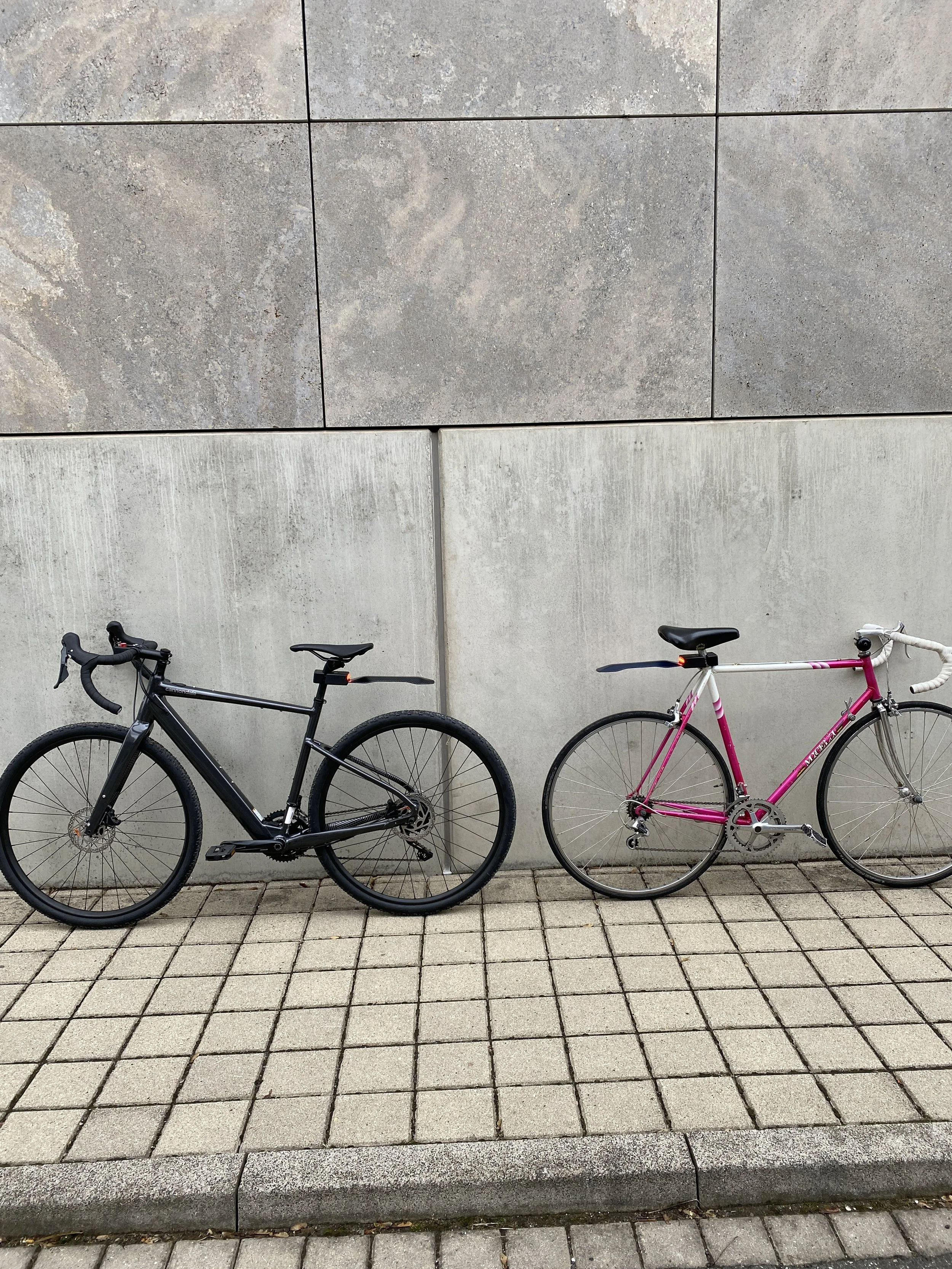 Two bicycles, one black and one pink, leaning against a tiled wall on a paved sidewalk.