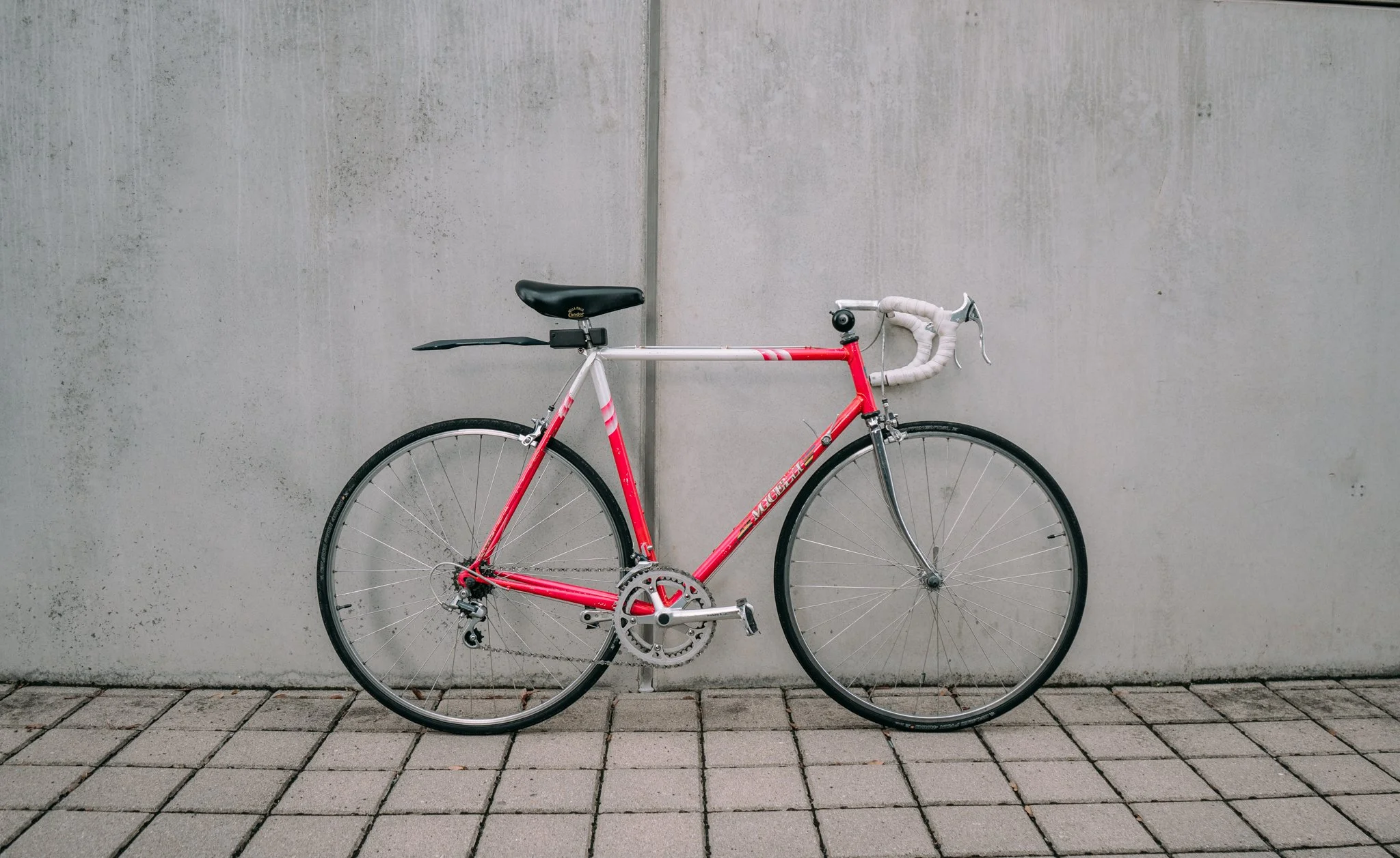 Red and white road bike with drop handlebars leaning against a concrete wall on a tiled surface.