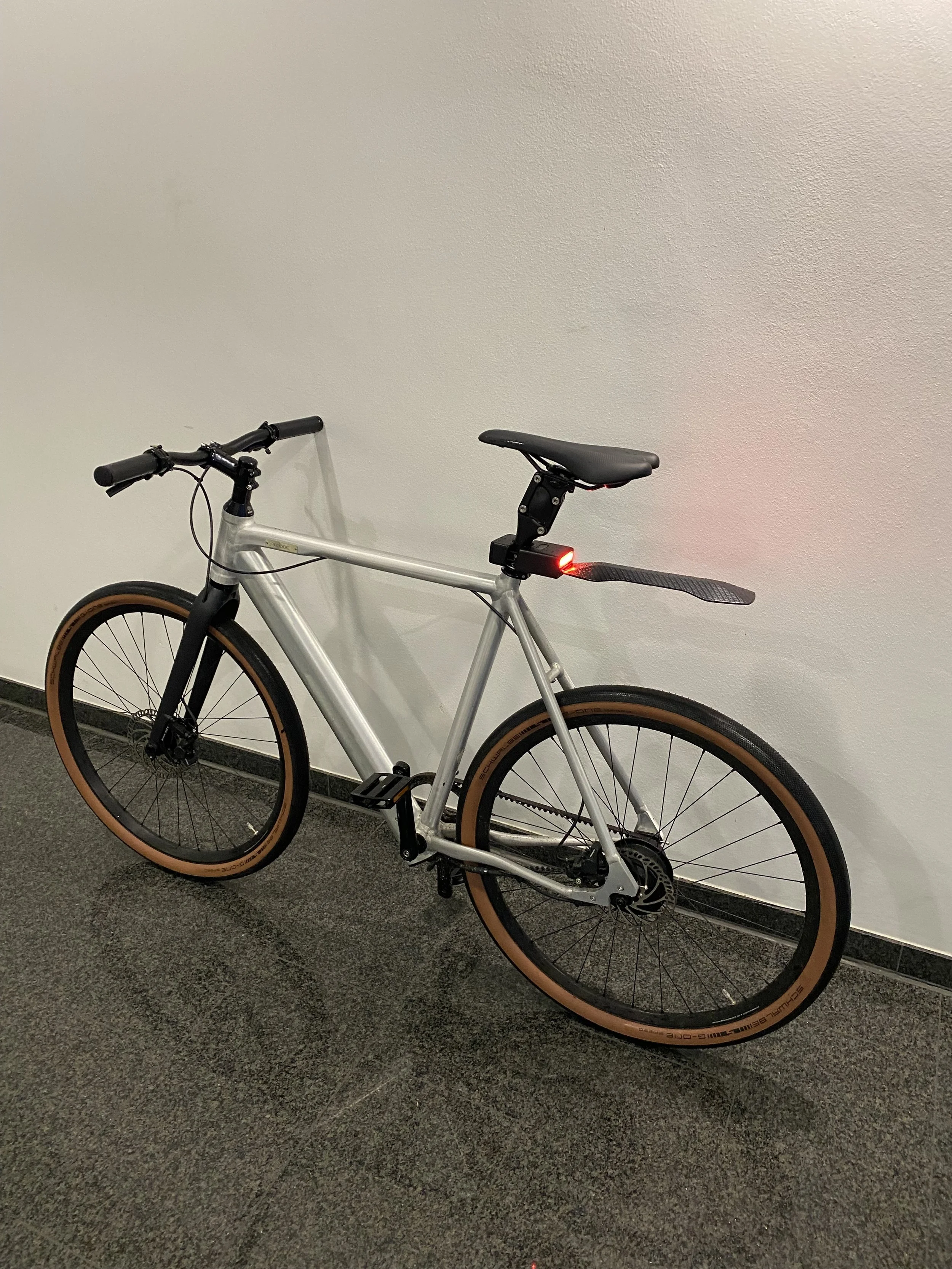 A sleek silver bicycle with brown tires, leaning against a white wall, featuring black handlebars and a small red light on the back.
