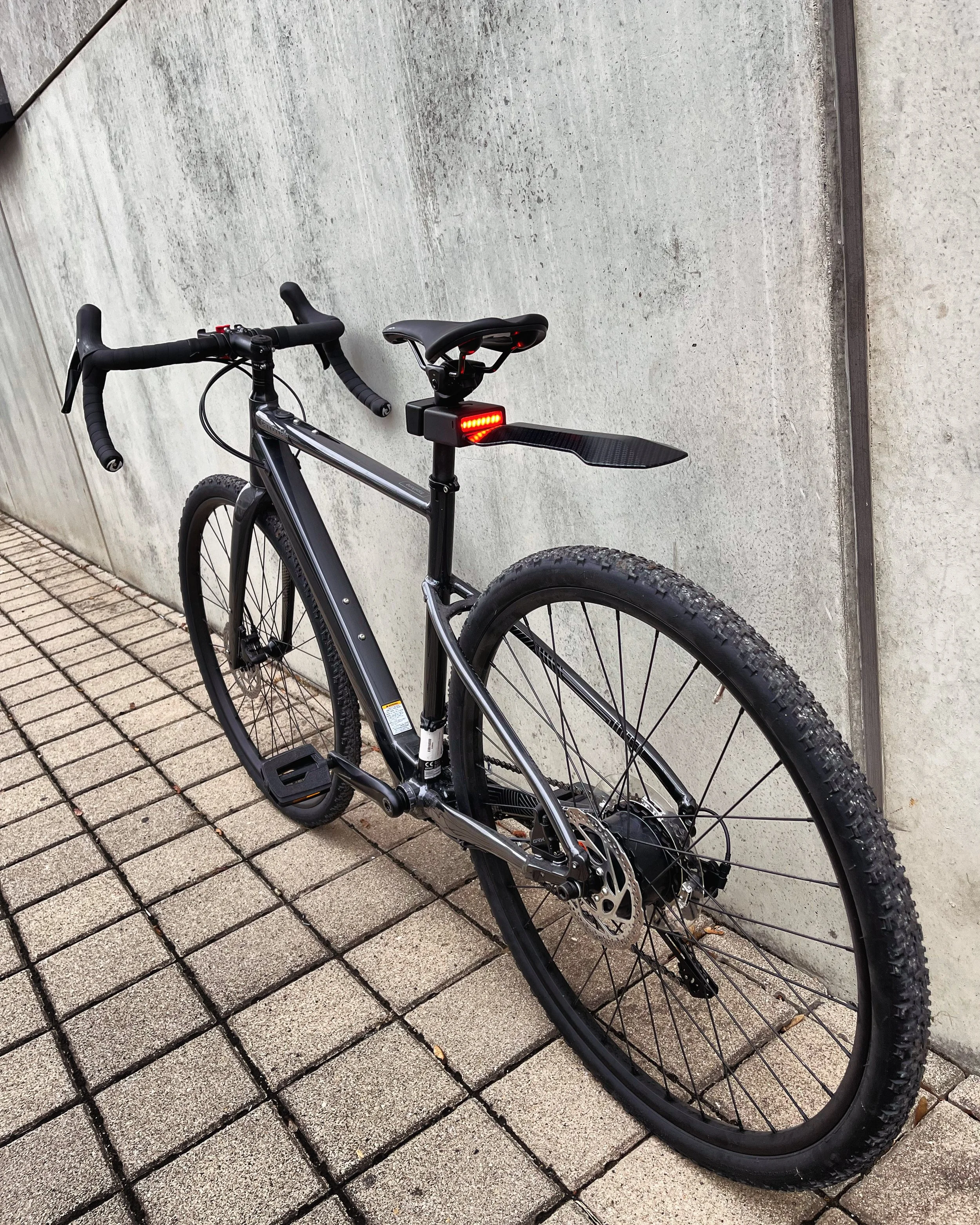 Black mountain bike with drop handlebars and rear mudguard, parked against a concrete wall on a tiled surface.
